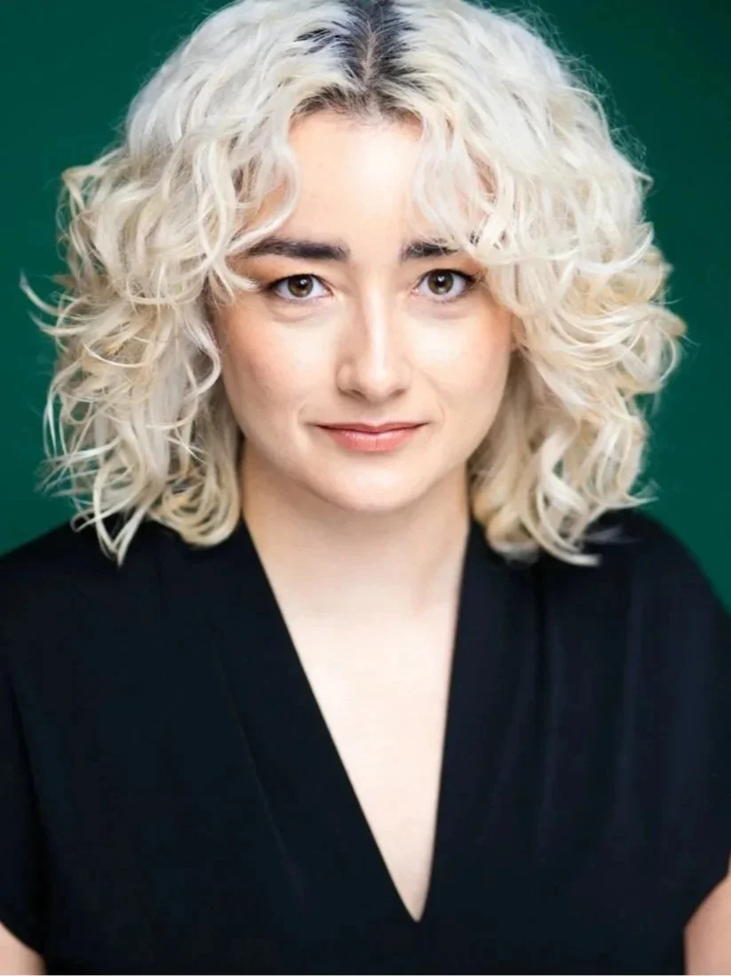 Carling is a white woman who is in front of a dark green background. She has curly blonde hair and dark eyebrows. She is smiling at the camera, wearing a deep v black top.