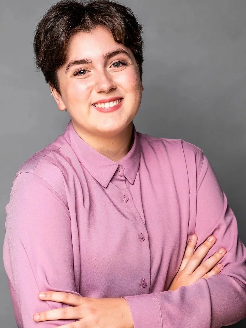Nell stands in front of a grey background. They have short dark hair and are smiling at the camera, with their arms crossed. They are wearing a pink button down shirt.