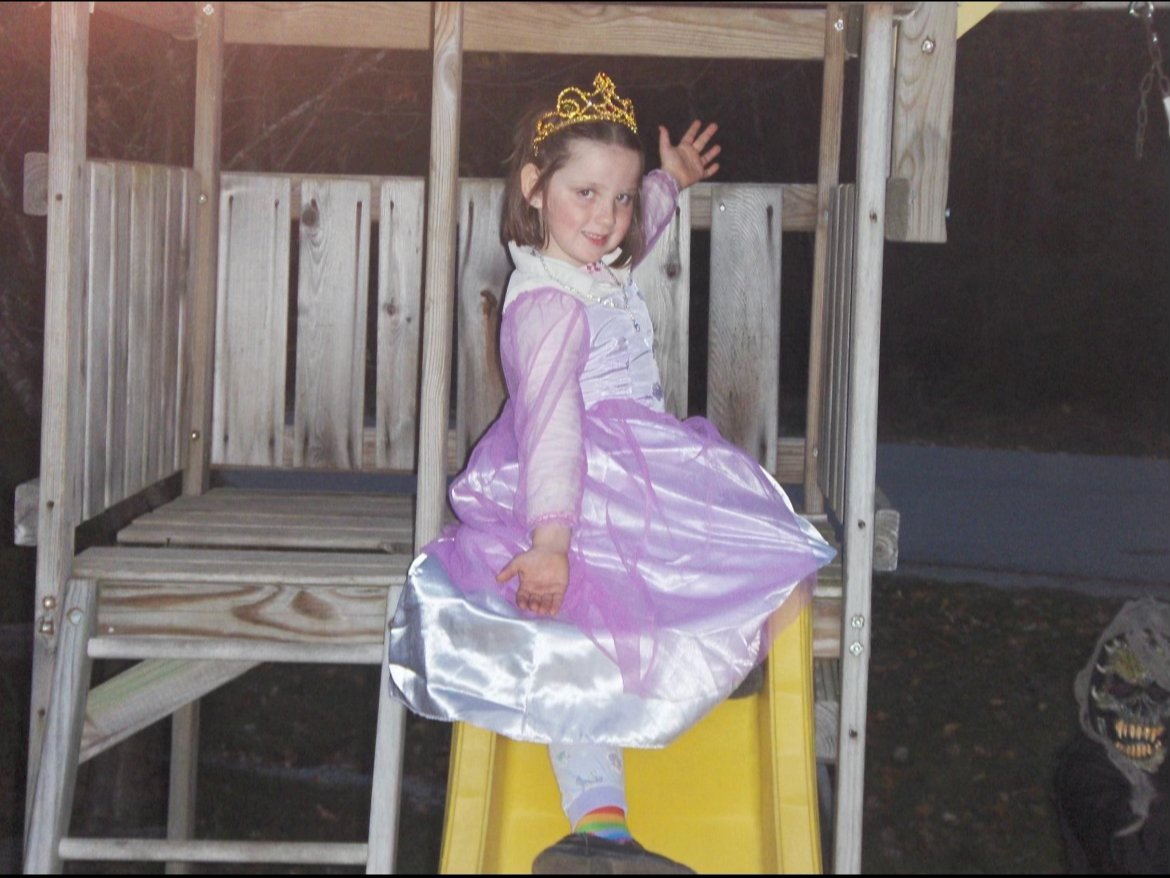 Little Grace poses at the top of a yellow slide, wearing a purple princess costume and a gold crown