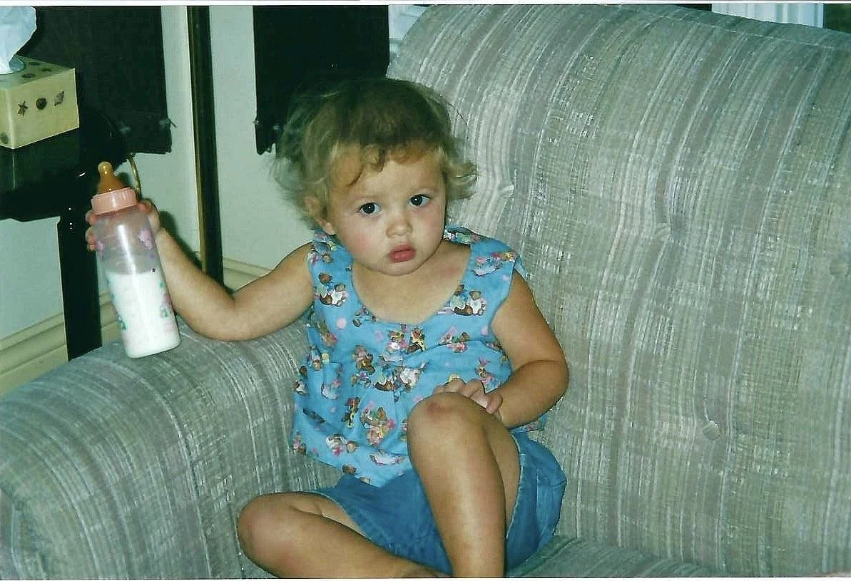 Little Meaghan sits on a grey couch, and looked at the camera with a pouty expression. She holds a half-full bottle in her hand, and is wearing a patterned blue shirt and denim shorts.
