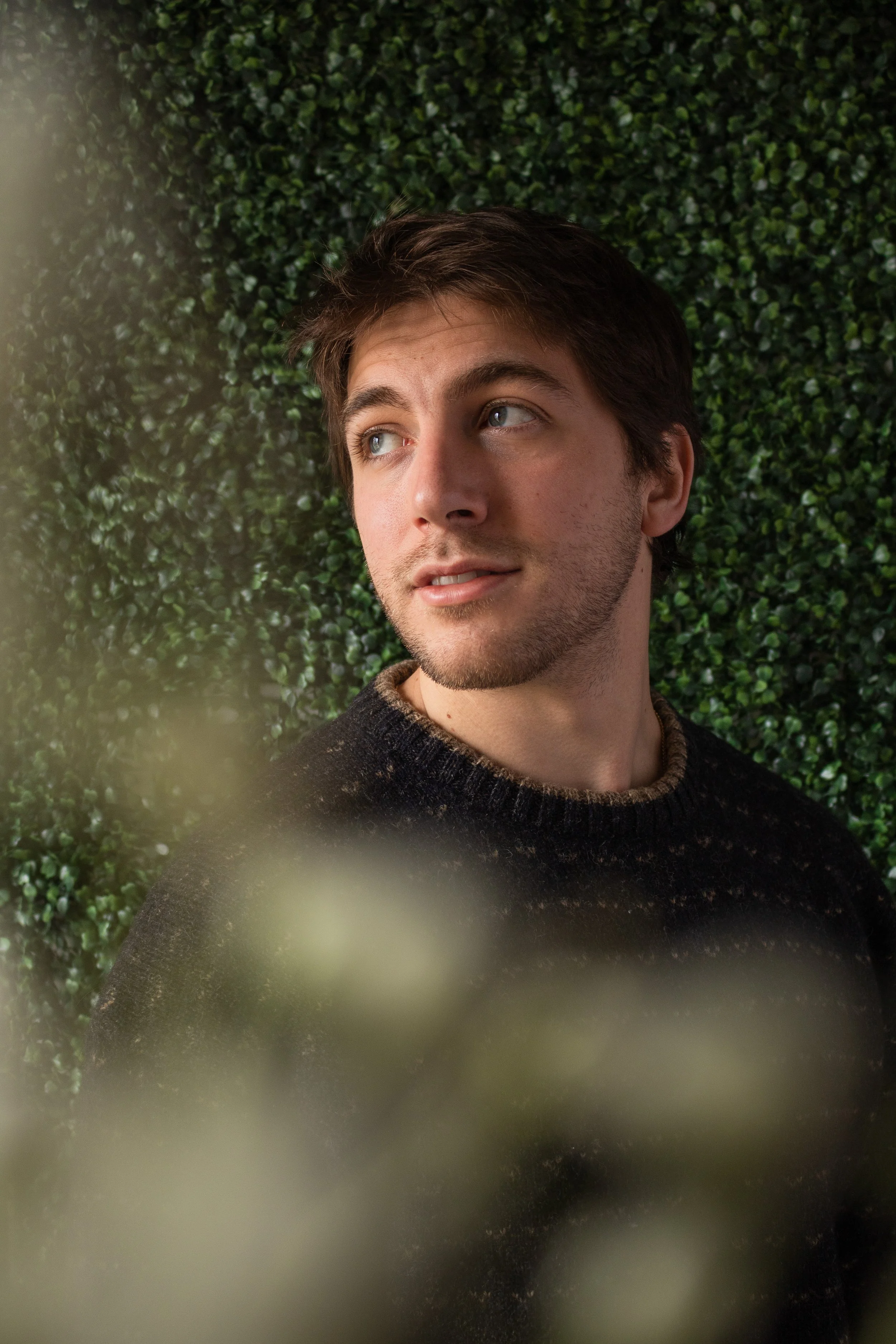 Lucas, a white man, looks off to the side of frame. He has short brown hair and a beard. He is wearing a knit dark sweater. Around the edge of frame are blurry white flowers, the background is green leaves.