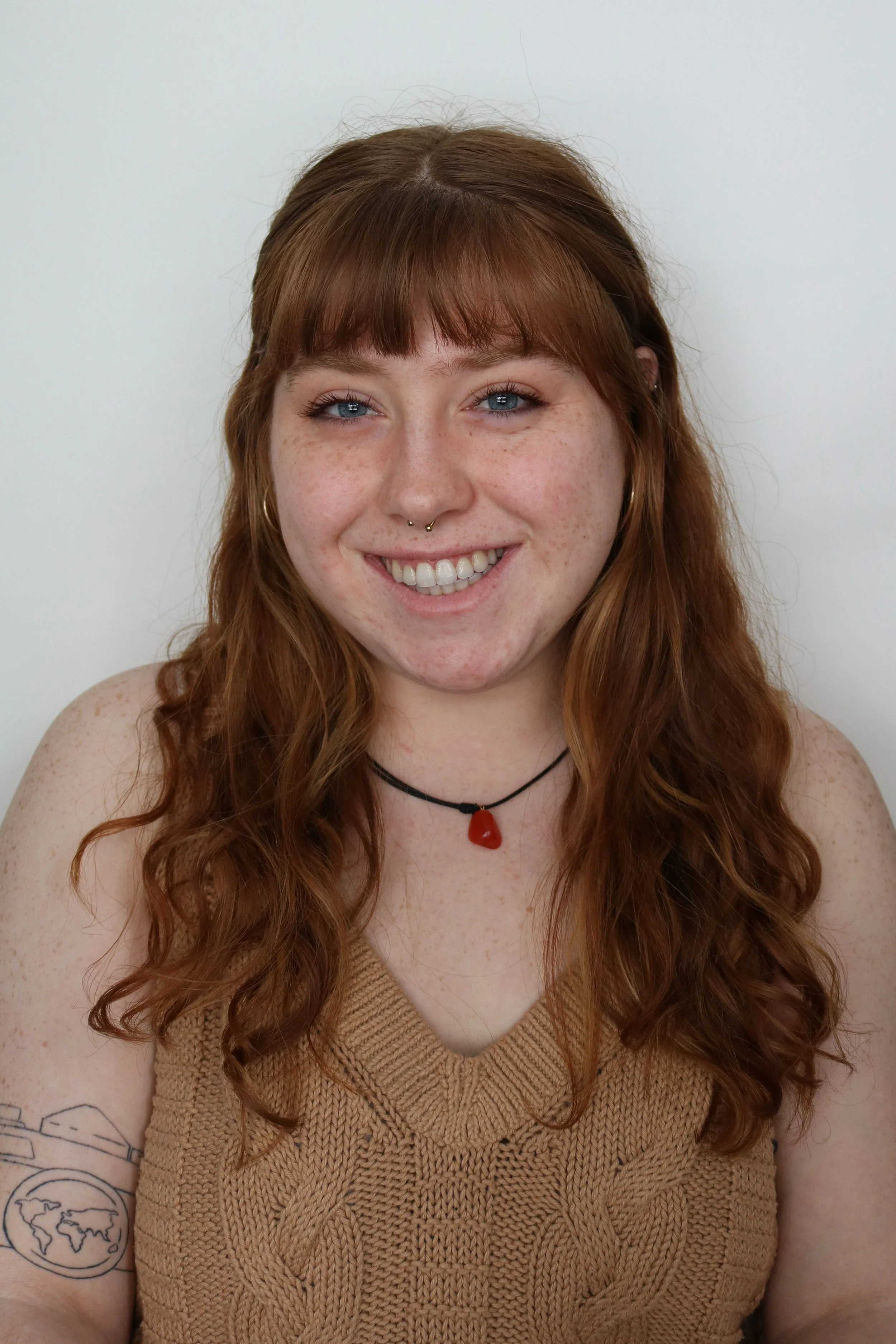 Chloe, a white woman, is smiling at the camera. She has long red hair and bangs, and is wearing a brown knit vest. The background is white.