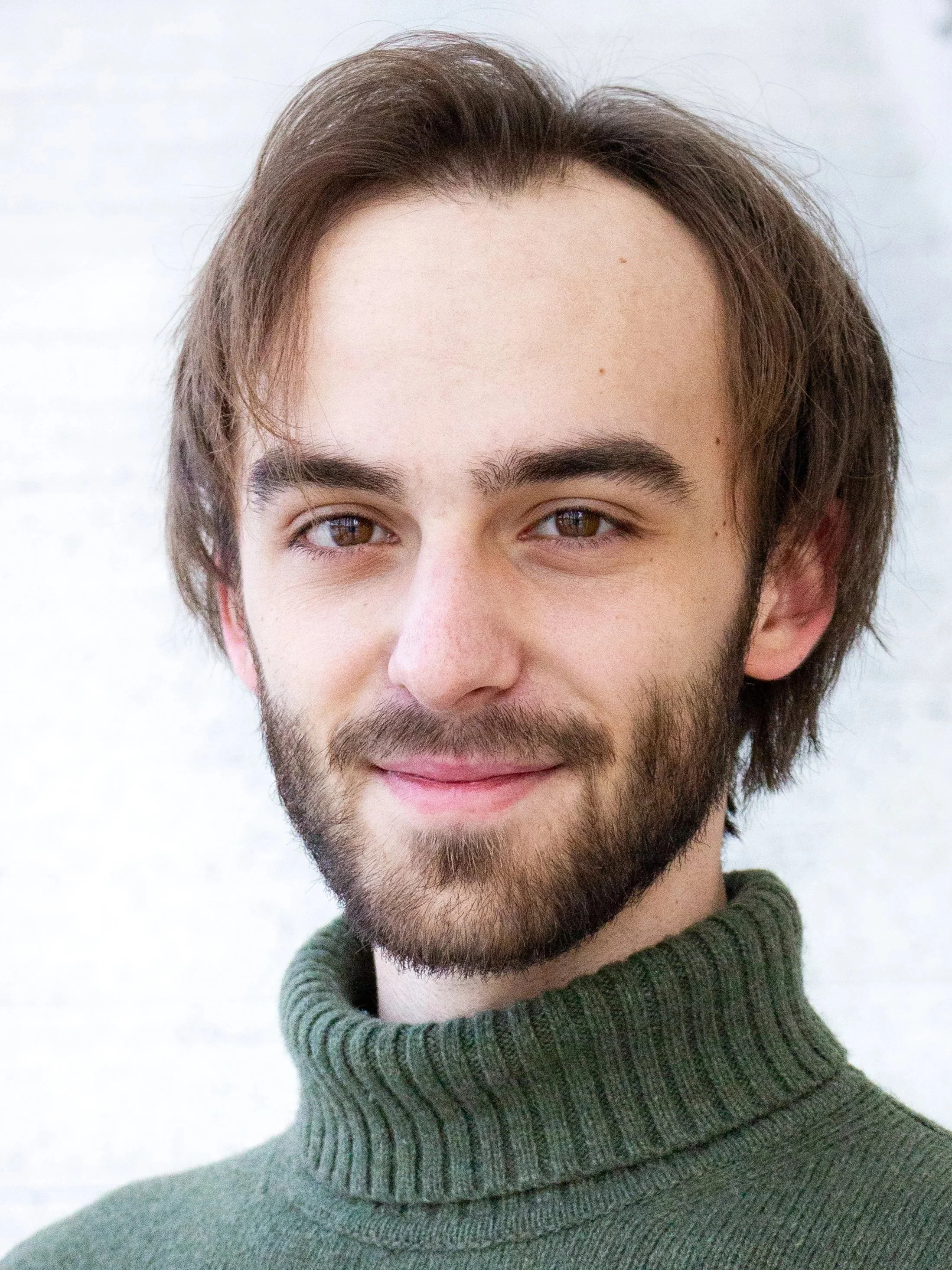 Mason, a white man, is looking to camera. He has brown hair and a beard, and is smiling. He is wearing a thick green turtleneck, and is in front of a white background.