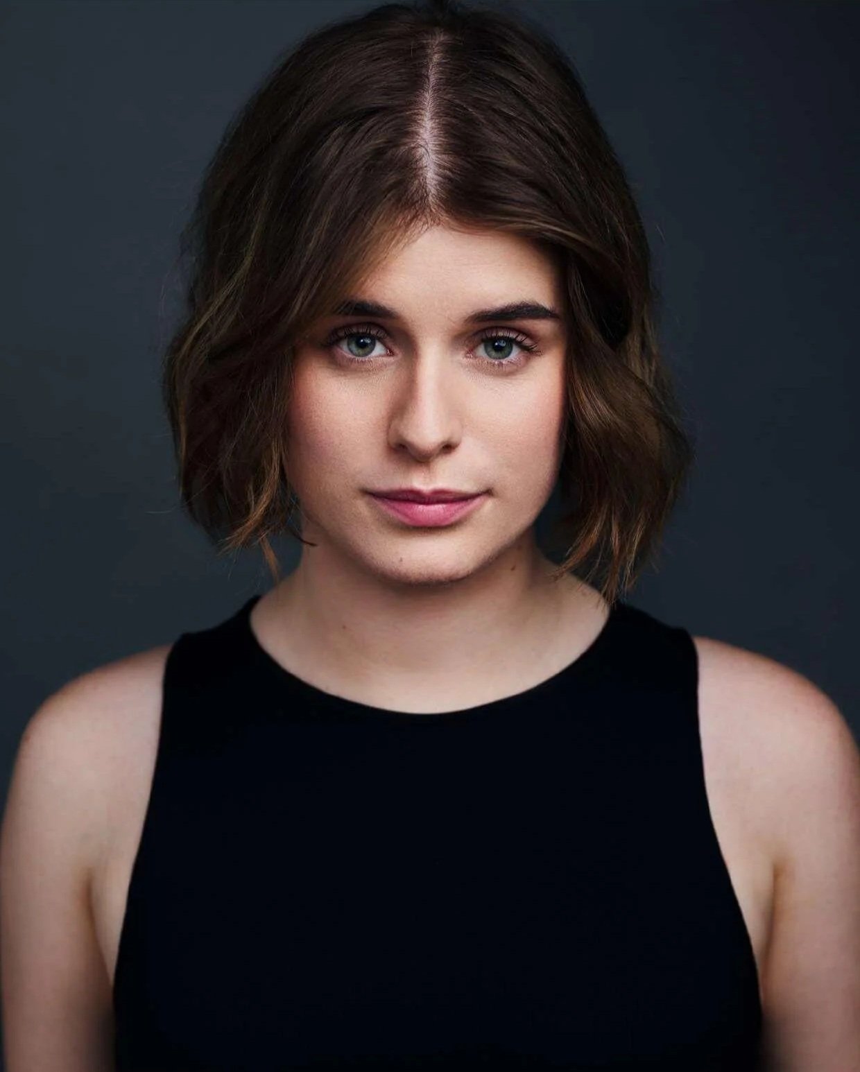 Sarah is a white woman with a dark brown bob. She is staring down the camera in front of a dark blue backgorund. She is wearing a black tank top.