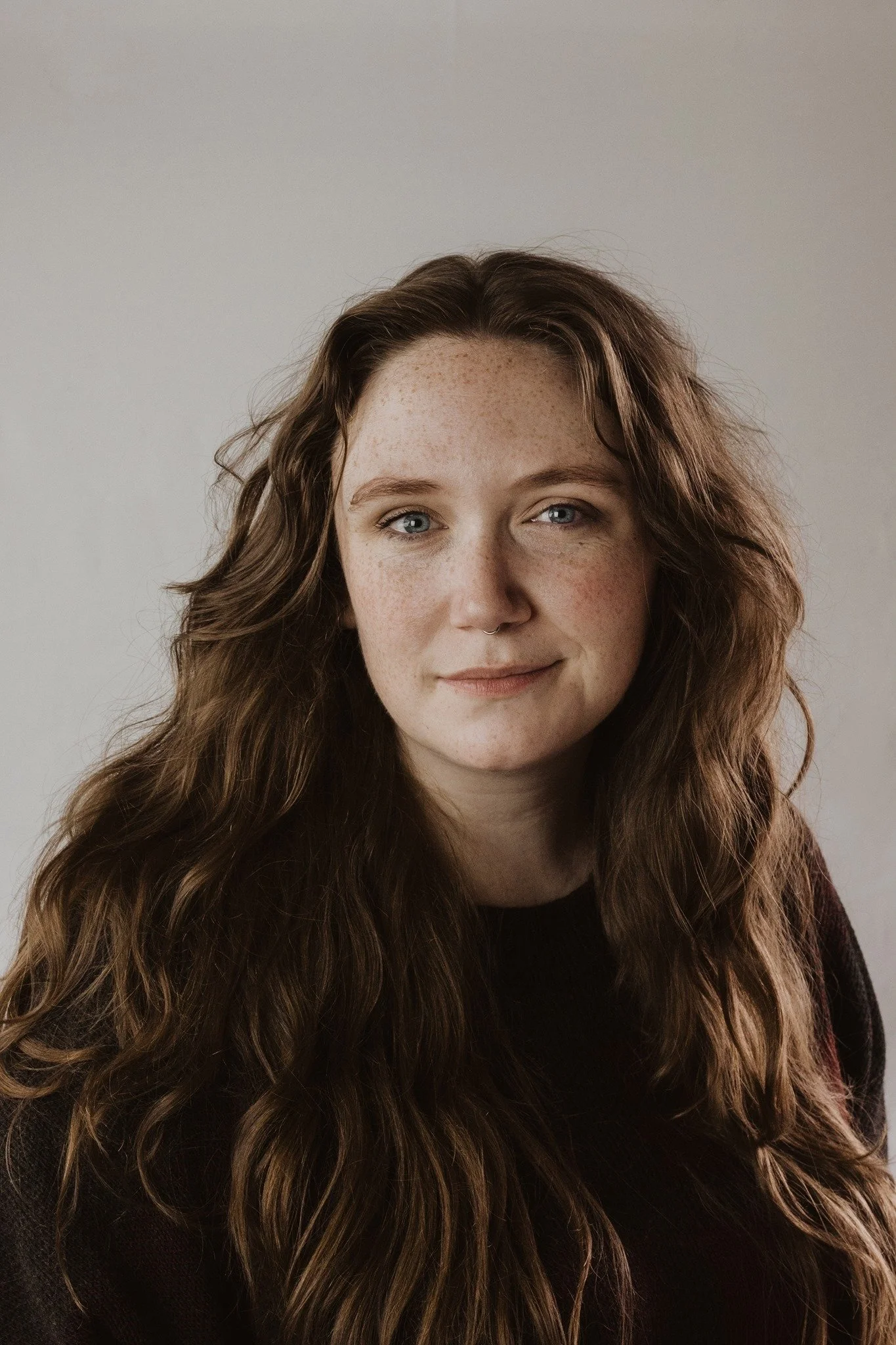 Rebecca, a white woman, looks at the camera with a soft smile. She has long auburn hair that is wavy, and is wearing a black top.