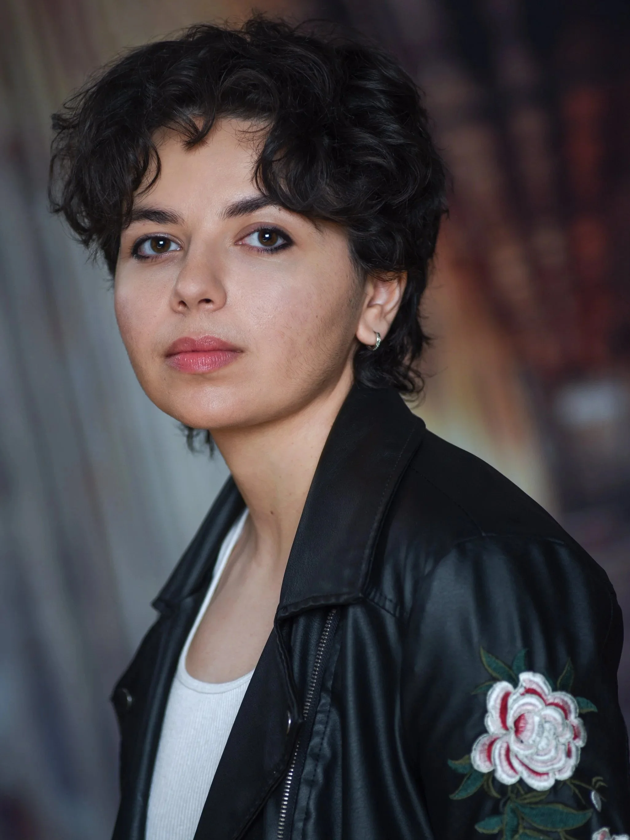 Jules is a nonbinary performer with warm skin. They have short black hair and are looking defiantly at the camera. They are wearing a black leather jacket with a rose embroidered on the sleeve