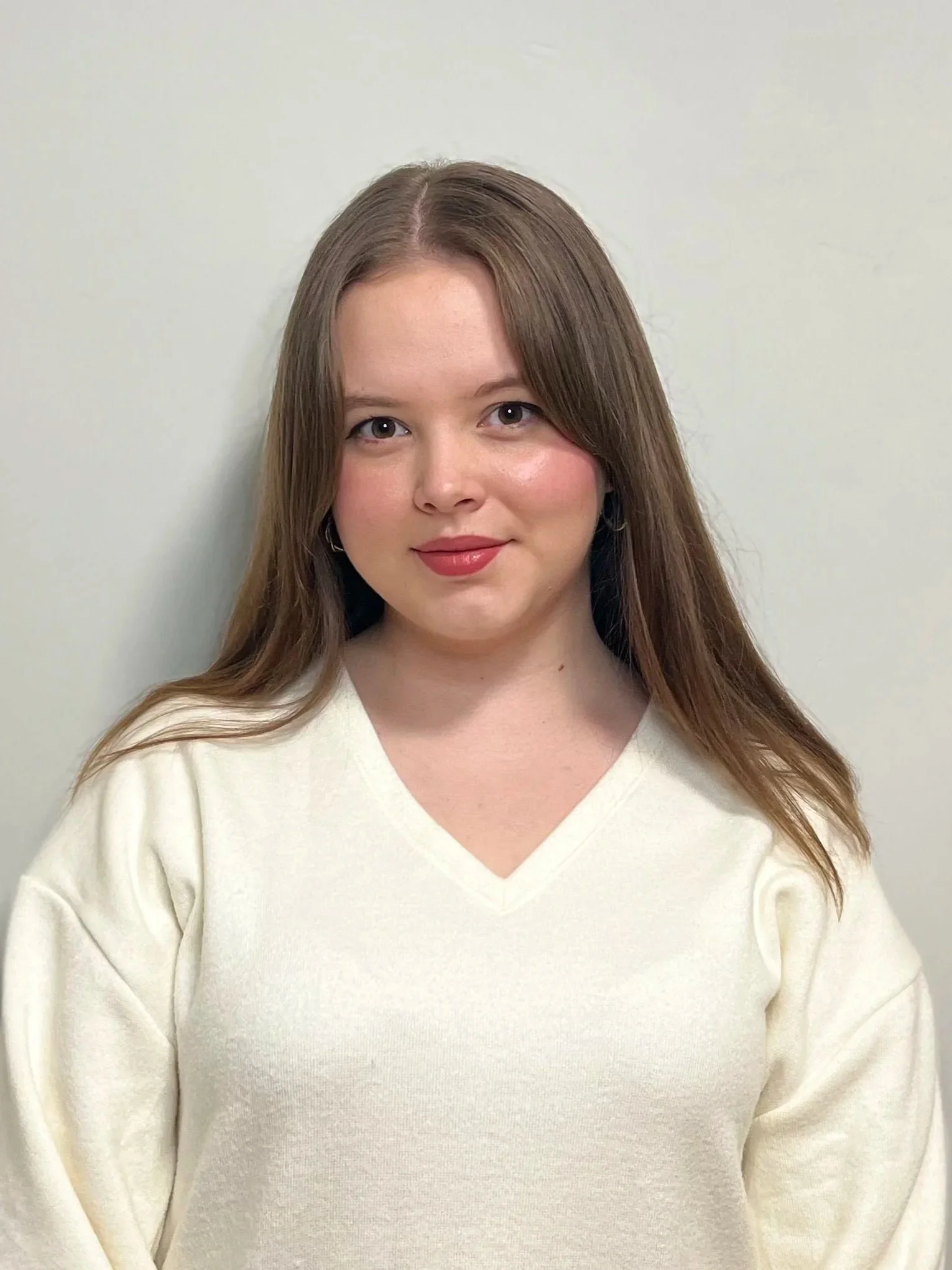 Meaghan, a white woman, stands in front of a light background. She has shoulder-length brown hair and is smiling at the camera. She is wearing a white v neck sweater.