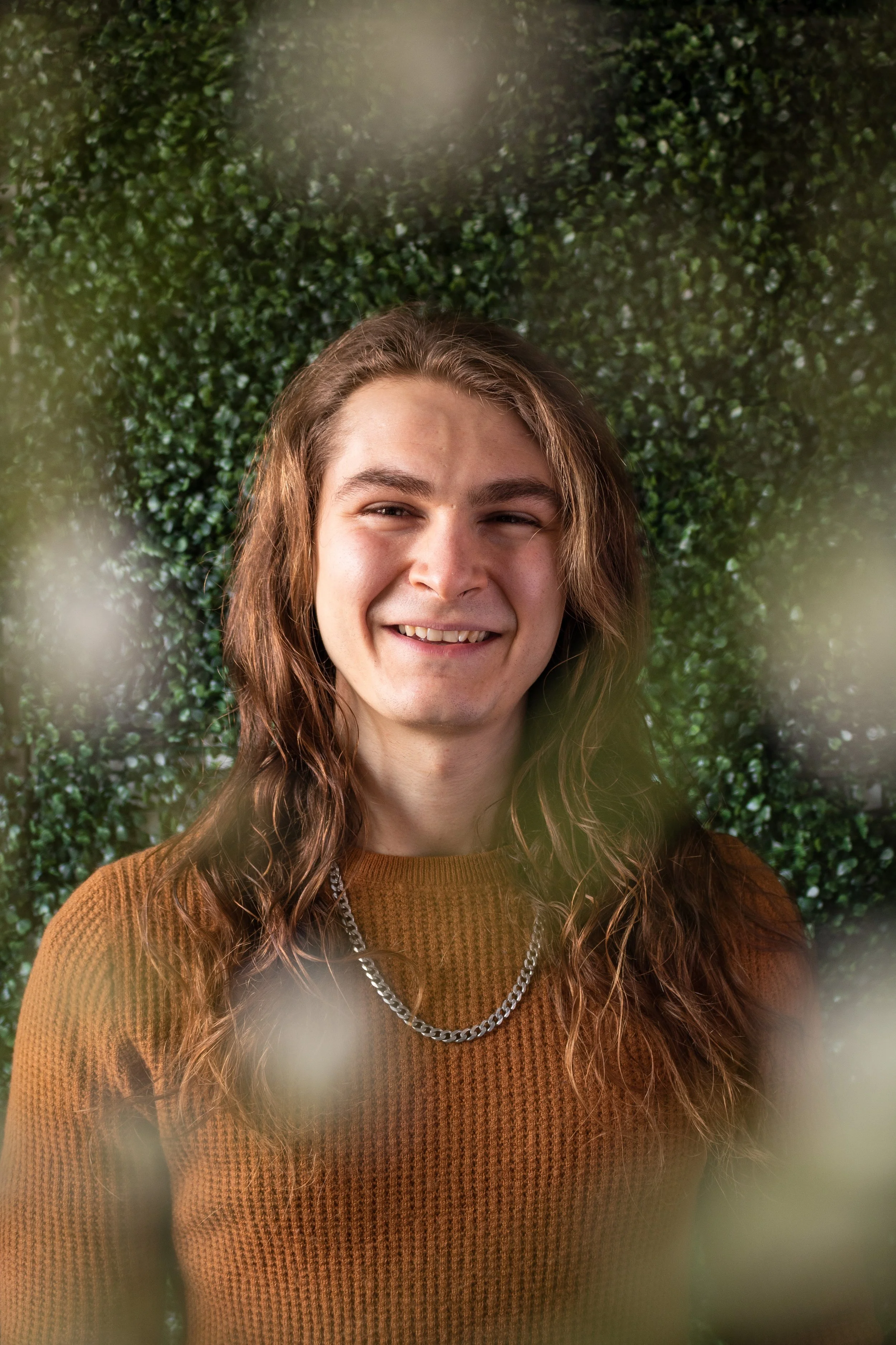 Blake, a white man, is smiling at the camera. He has long brown hair, and a goatee. He is wearing a pumpkin sweater and silver chain. Around the edge of frame are blurry white flowers, the background is green leaves.