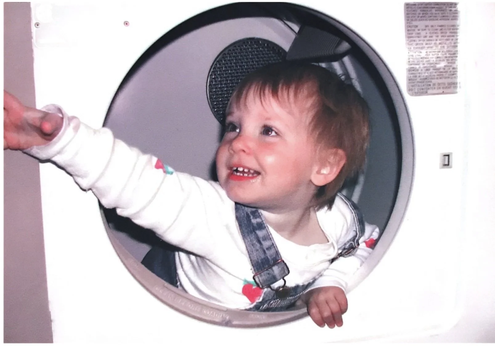 Little Annie is in the drum of a front-loading dryer, reaching to the side of frame. She is smiling, and wearing a long sleeve white shirt and denim overalls.