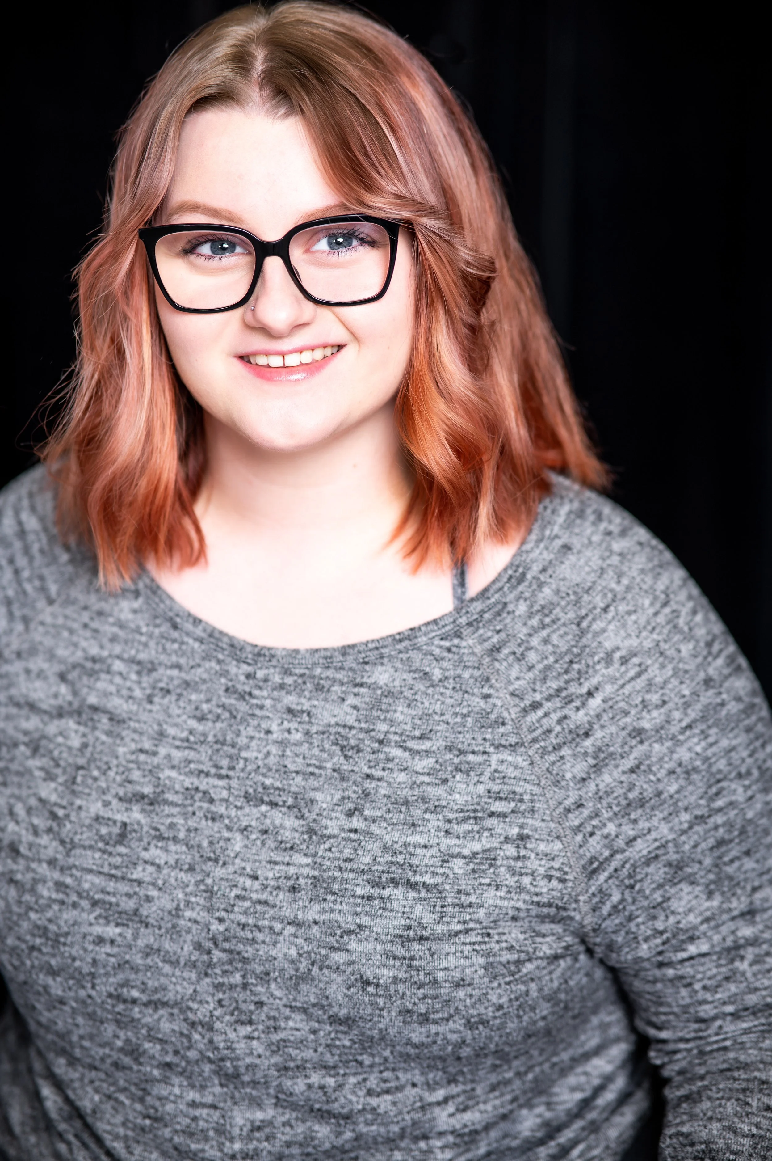 Emilia, a white person, is smiling at the camera. They have shoulder-length pink hair, and glasses. She is wearing a grey blouse. The background is black.