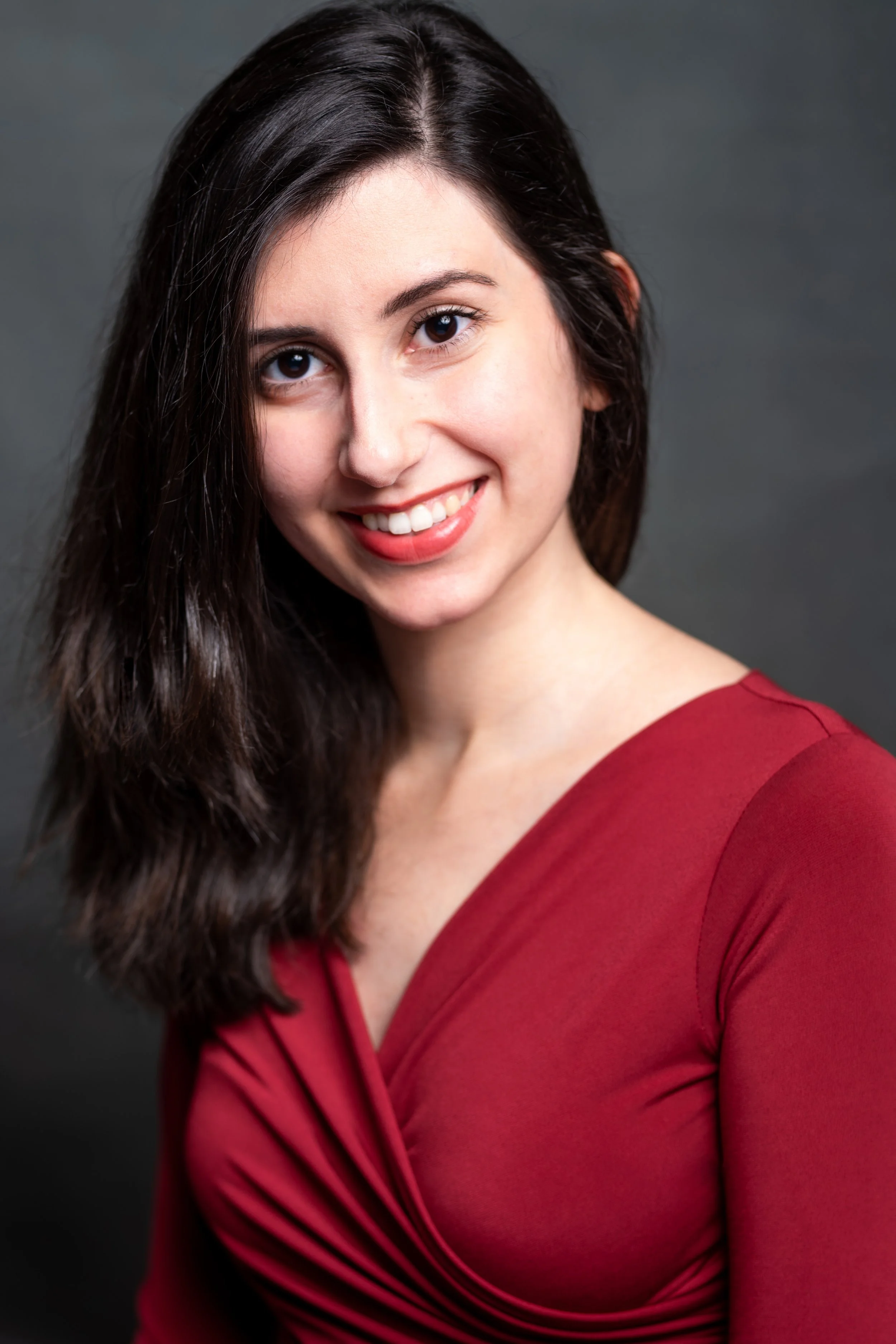 Elif, a Turkish-Canadian woman, is smiling at the camera. She has medium length black hair, and is wearing a red blouse. The background is grey.