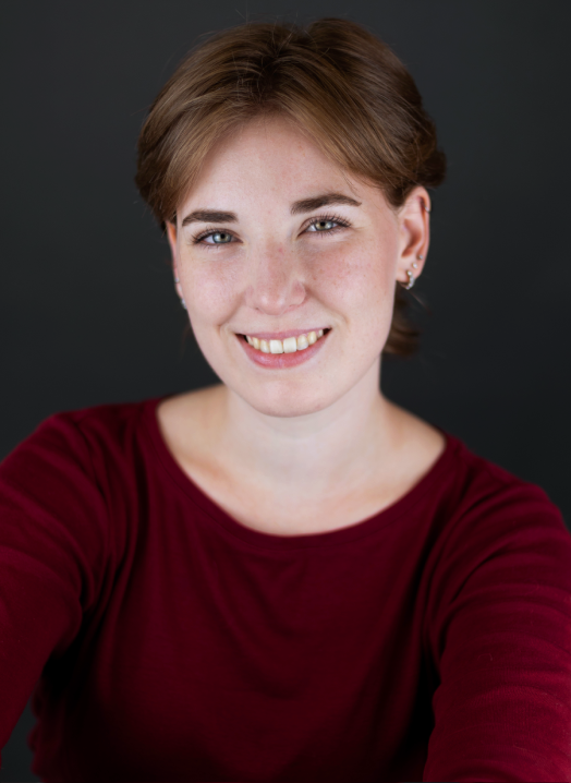 Annie, a white person, smiles at the camera. They have short brown hair and is wearing a red blouse. The background is dark grey.