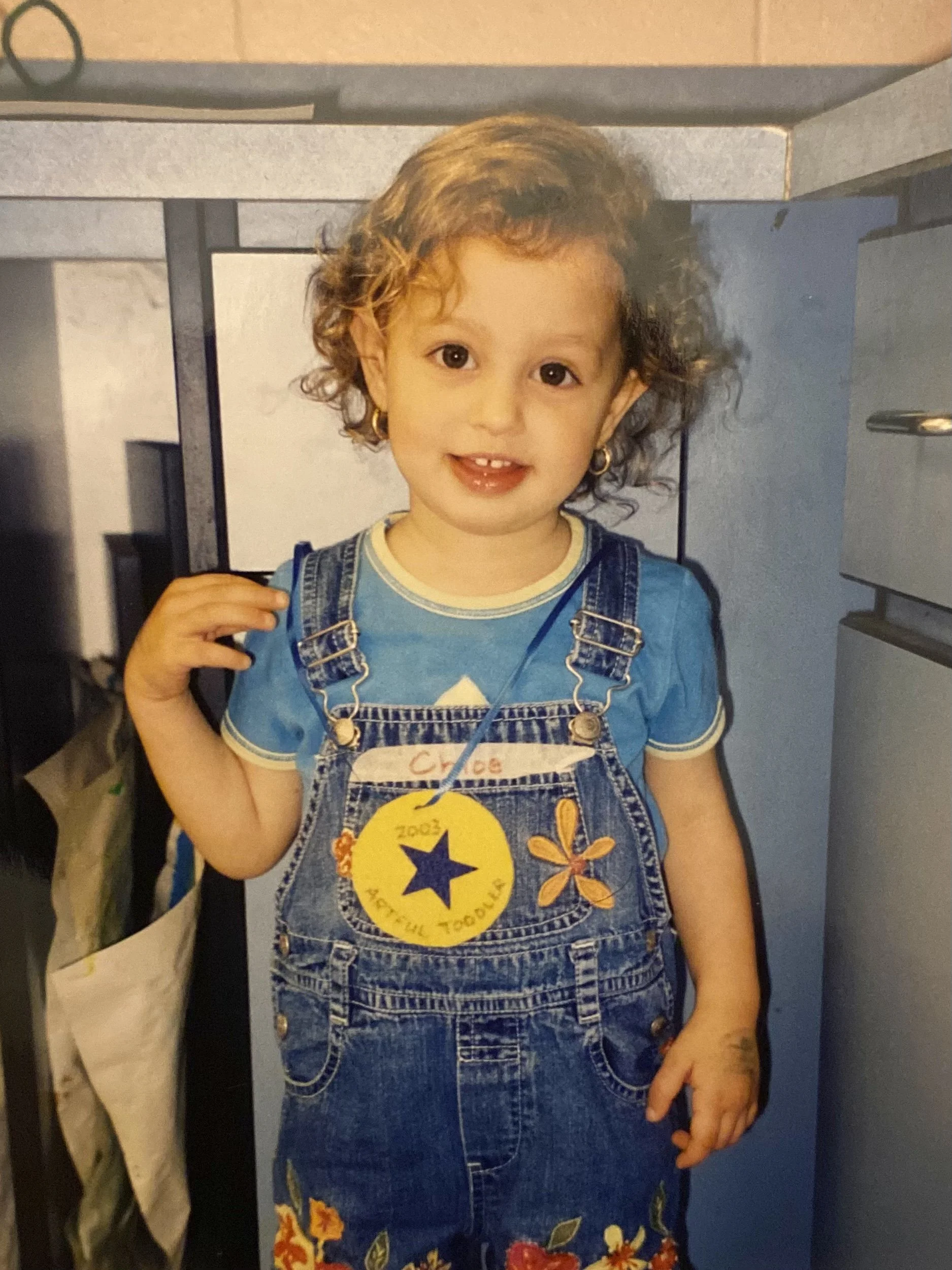 Little Chloe smiling at the camera. She is wearing overalls, and has a paper award star around her neck.