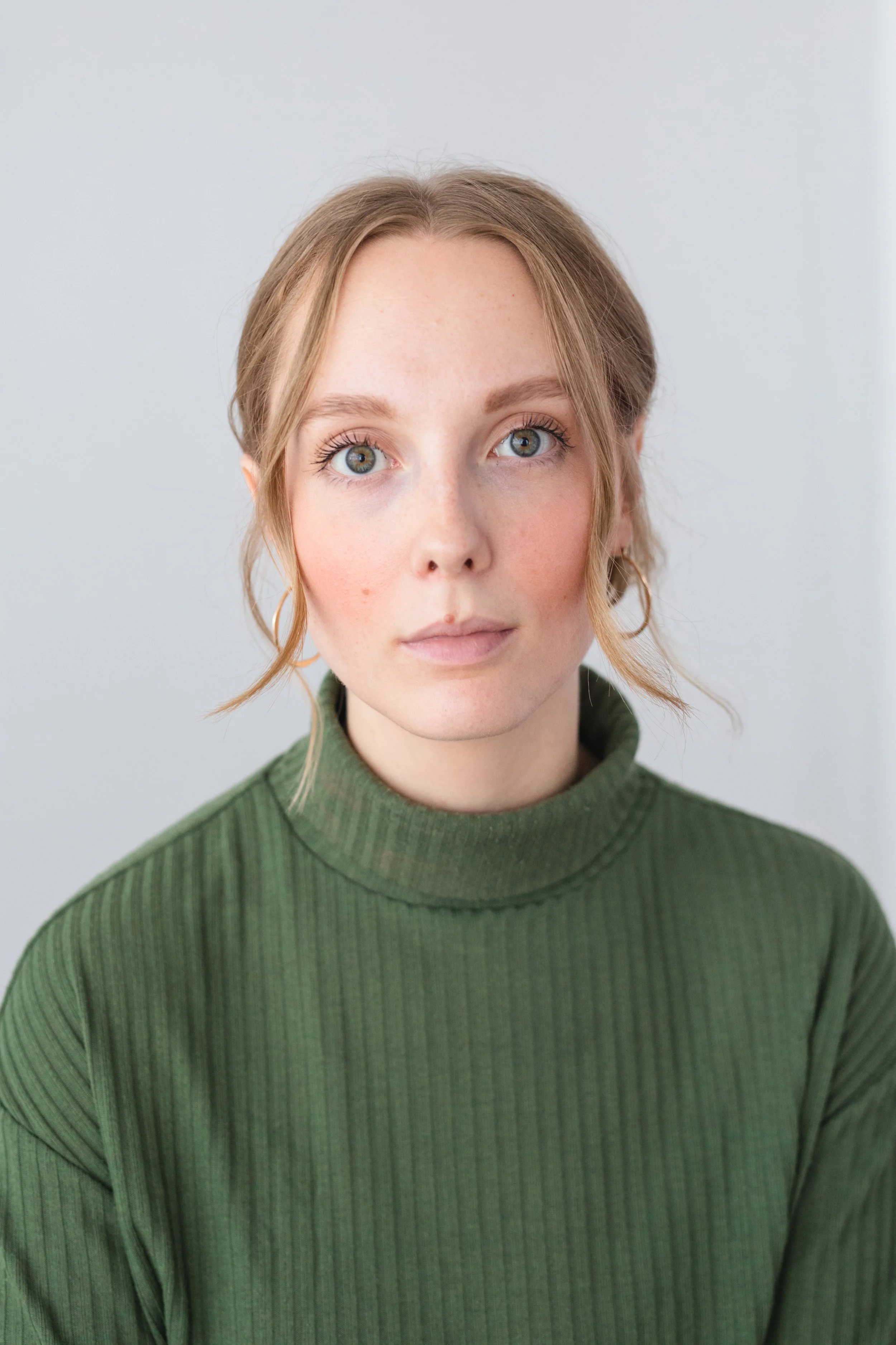 Rachel is a white woman with blonde hair and green eyes, which match her green turtleneck. She is looking softly to camera, in front of a white background.
