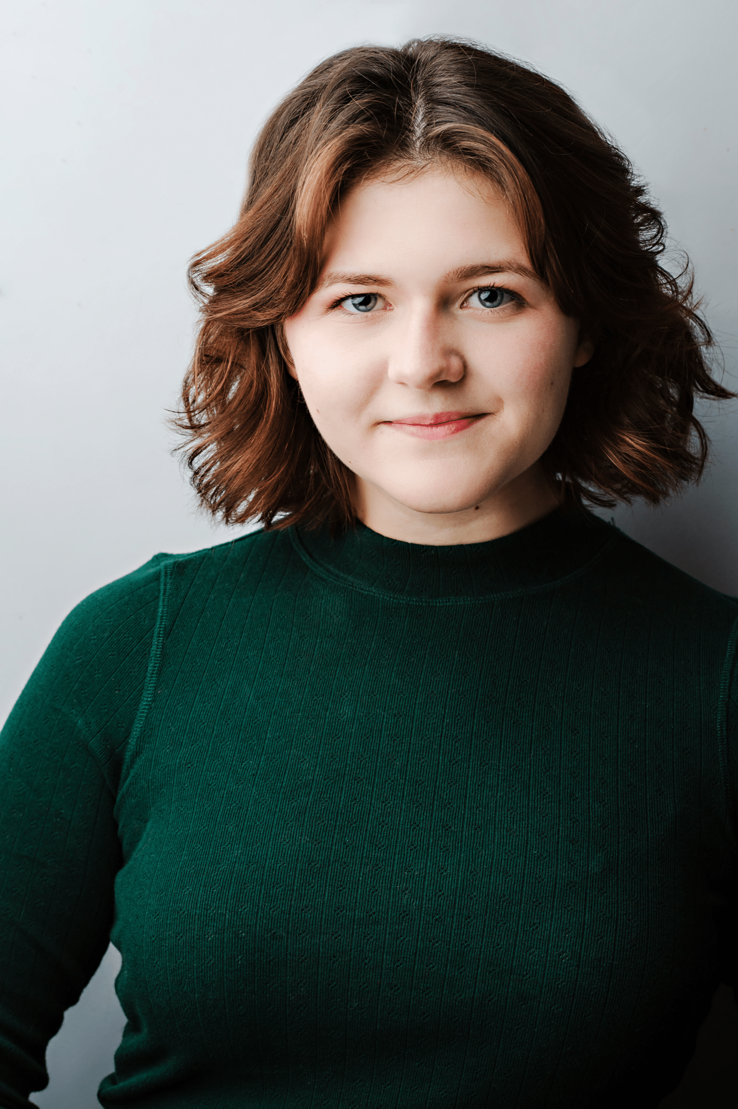Cheyenne, a white woman, stands in front of a white background. She has a light brown bob and blue eyes. She has a small smile, and is wearing a dark green turtleneck