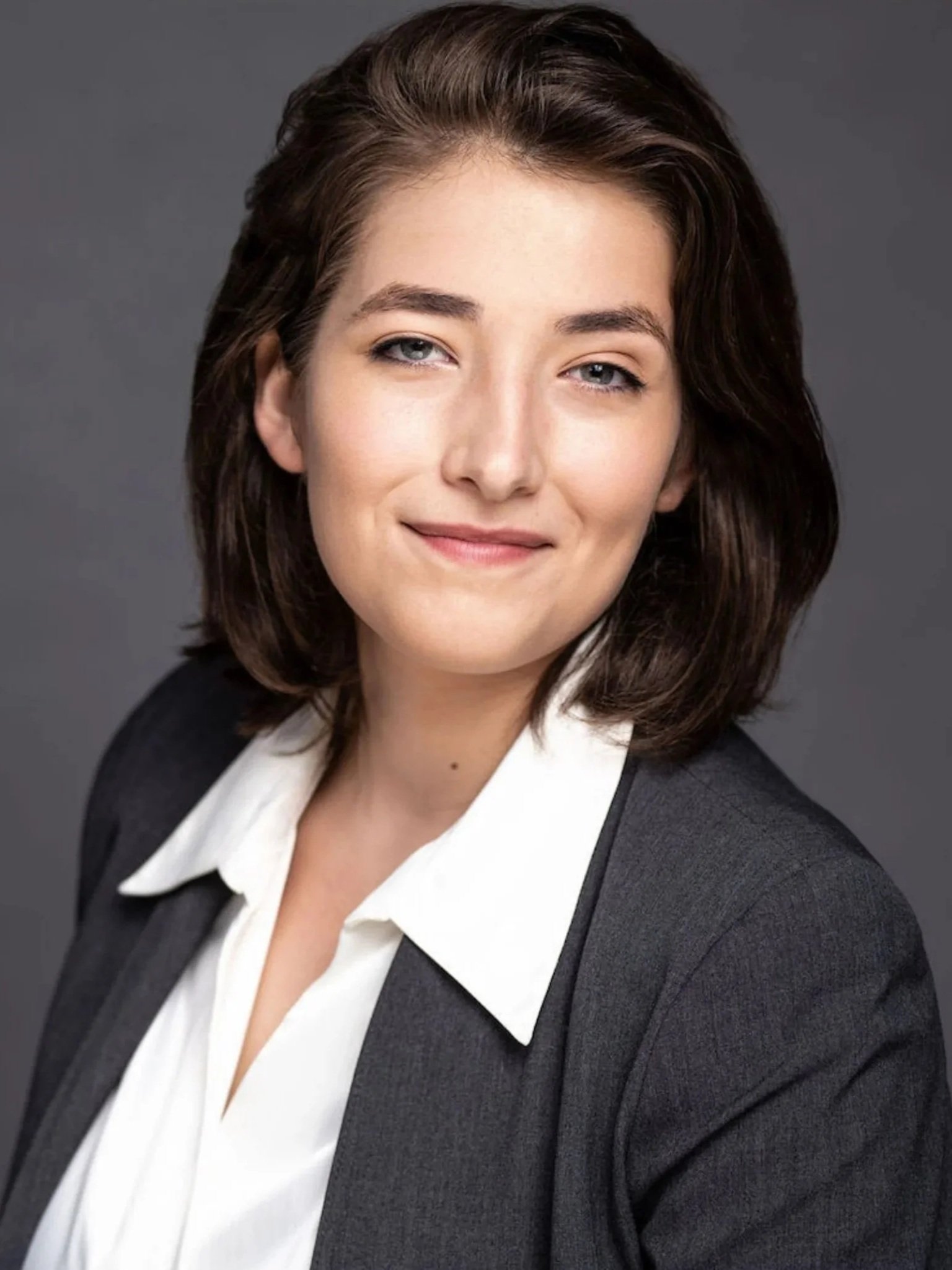 Saskia, a white woman, has shoulder length brown hair that is swoopeed back from her face. She is smiling at the camera. ?She is wearing a white collared shirt and grey blazer, which matches the grey background behind her.