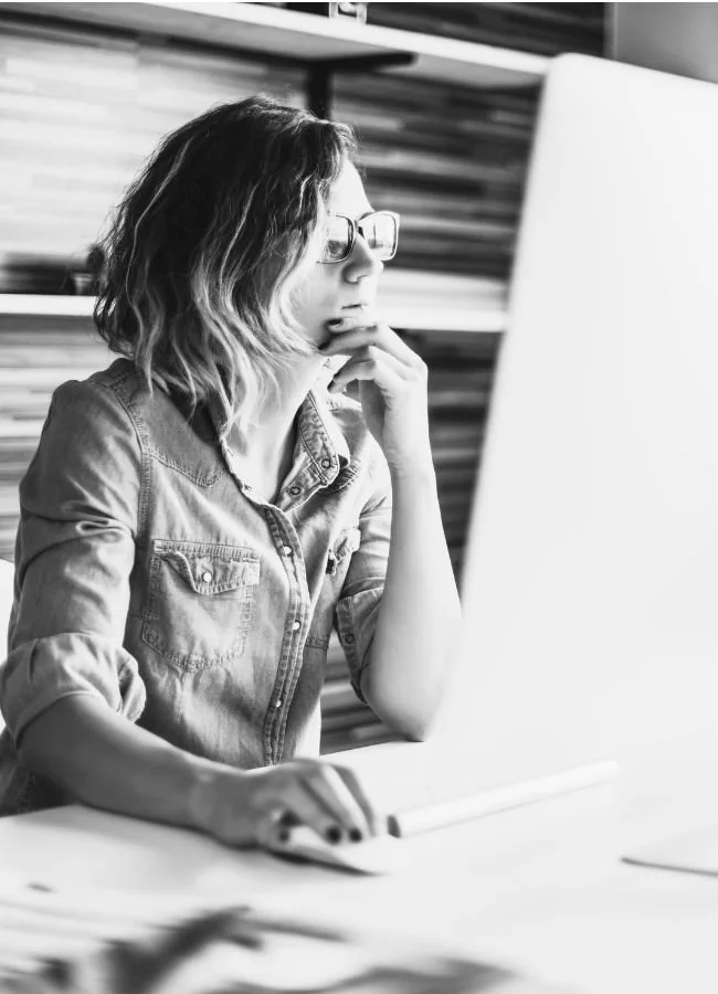 A woman with shoulder-length wavy hair and glasses working on a computer in an office setting.