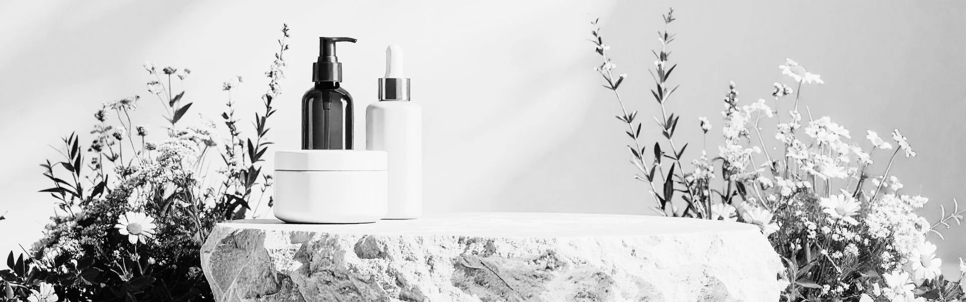Black and white photo of skincare or cosmetic bottles and containers on a rough, stone surface, surrounded by flowers and foliage.