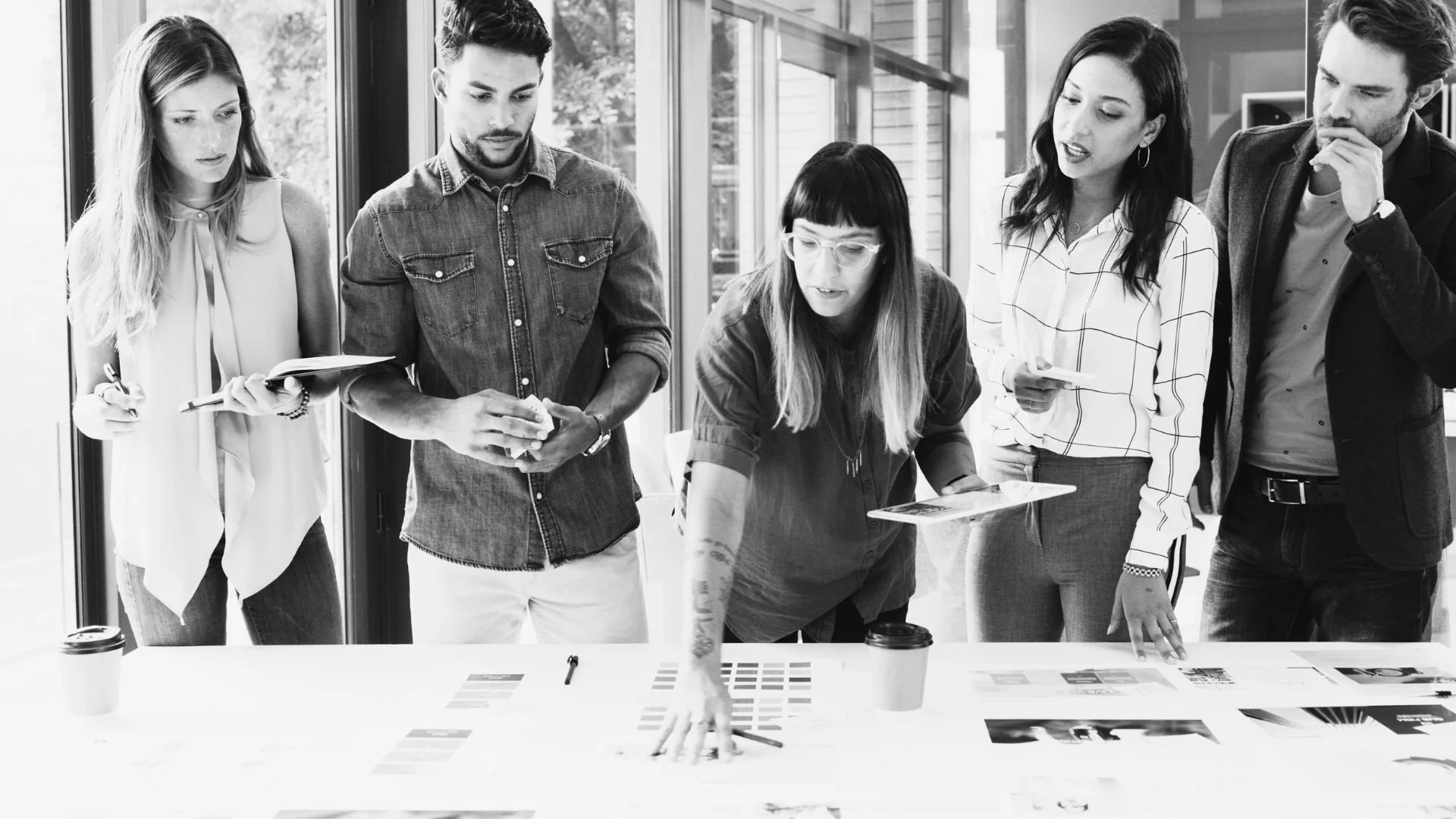 Five people gathered around a table in a modern office, engaged in a discussion or presentation, with charts and papers spread on the table.