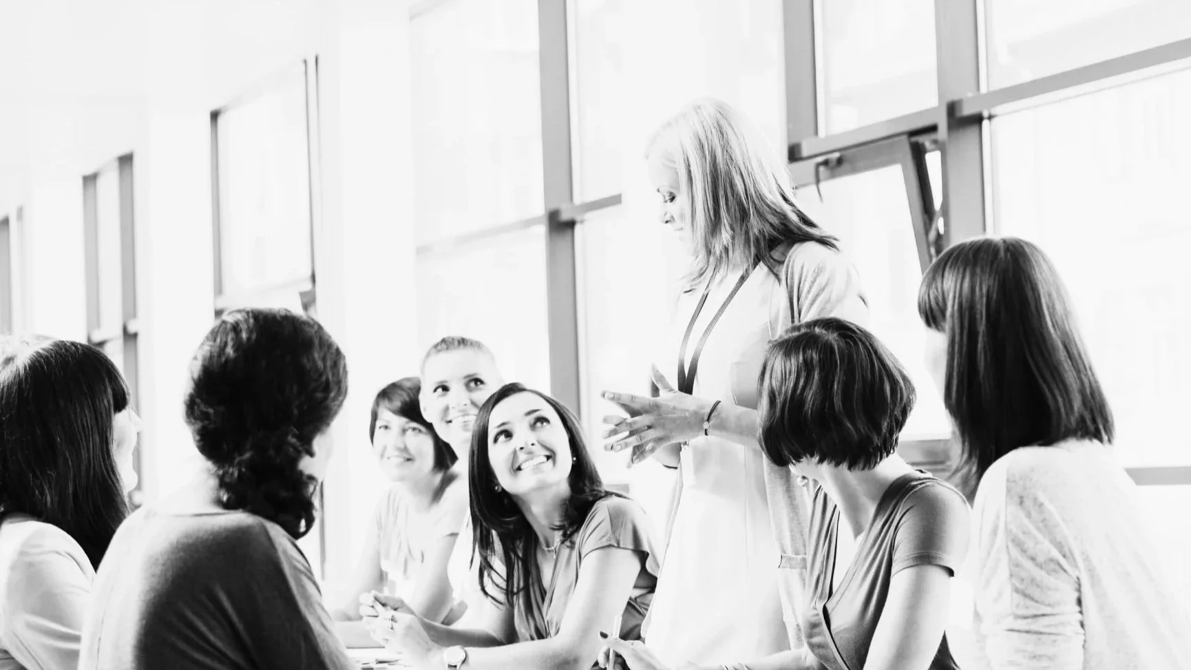 Group of women sitting at a table engaged in conversation, with one woman standing and speaking to the others, in a bright room with large windows.