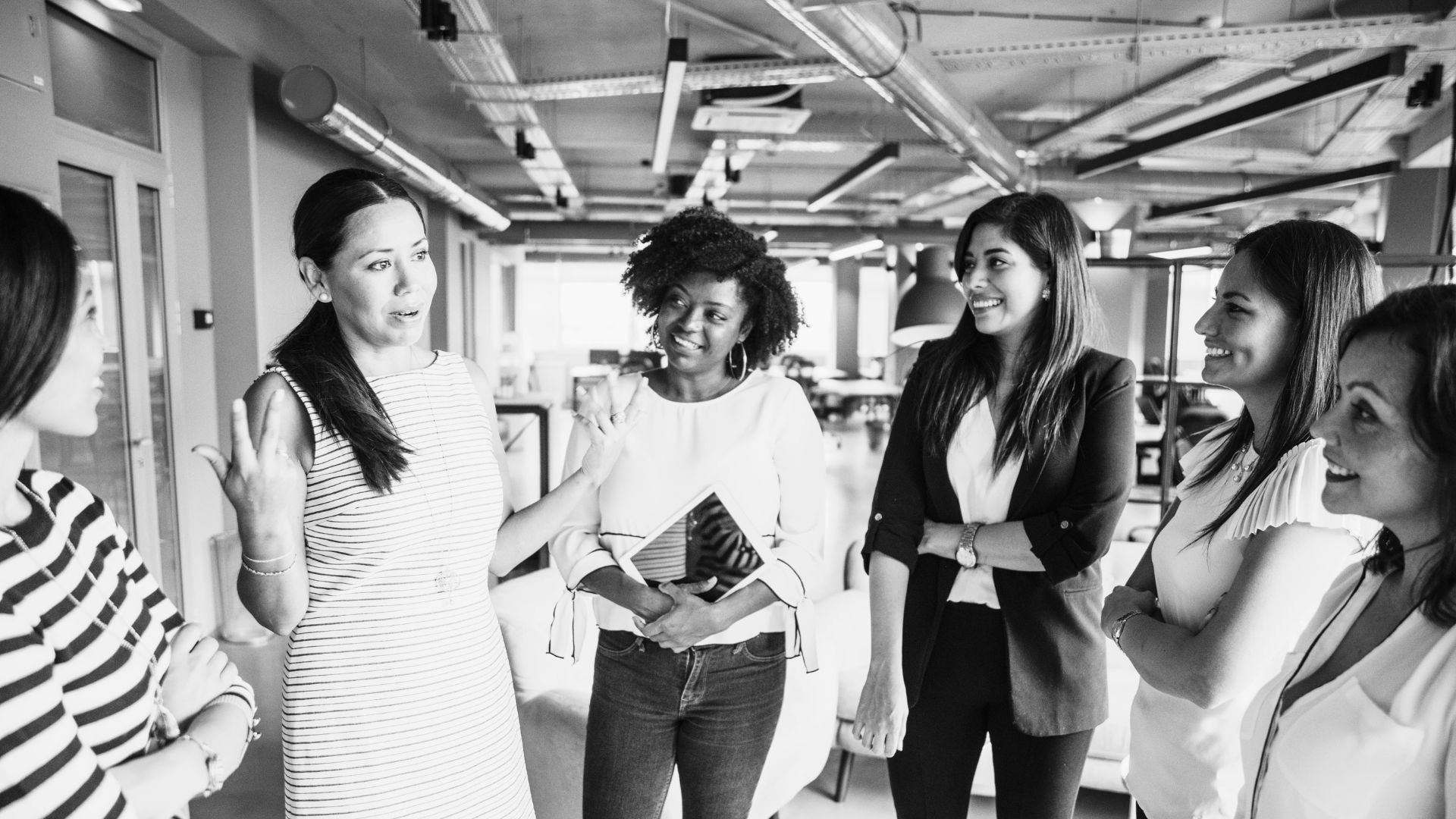 Six women having a conversation in an office setting, smiling and engaged with each other.