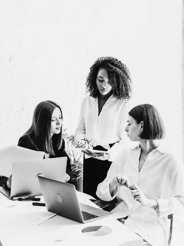 Three women in a meeting, two seated at a desk with laptops and one standing, holding a tablet and showing it to the others.