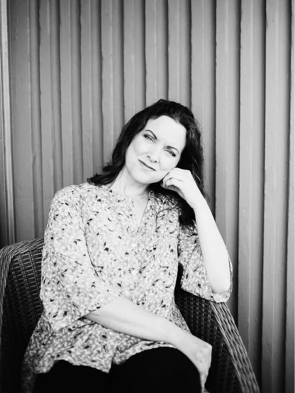 Kristy O'Leary sitting on a wicker chair in front of vertical wooden panels, smiling slightly and resting her head on her hand.
