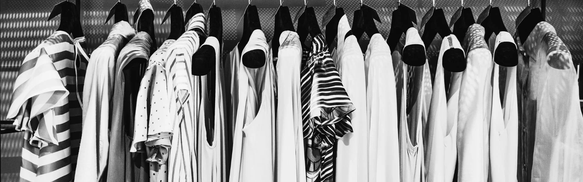 Black and white photo of various sleeveless tops, shirts, and dresses hanging on black hangers on a clothing rack.
