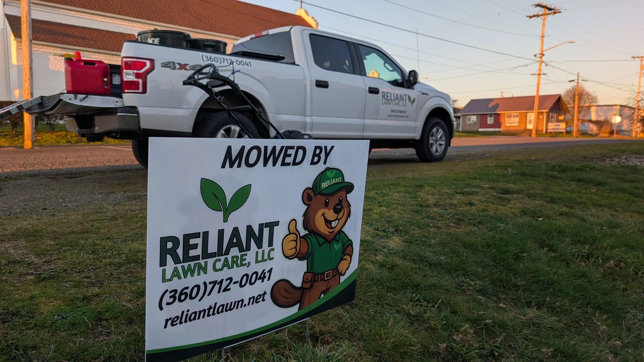 A Reliant Lawn Care LLC truck parked on a grassy area during sunset, with a sign in front of it showing the company's logo and contact information.