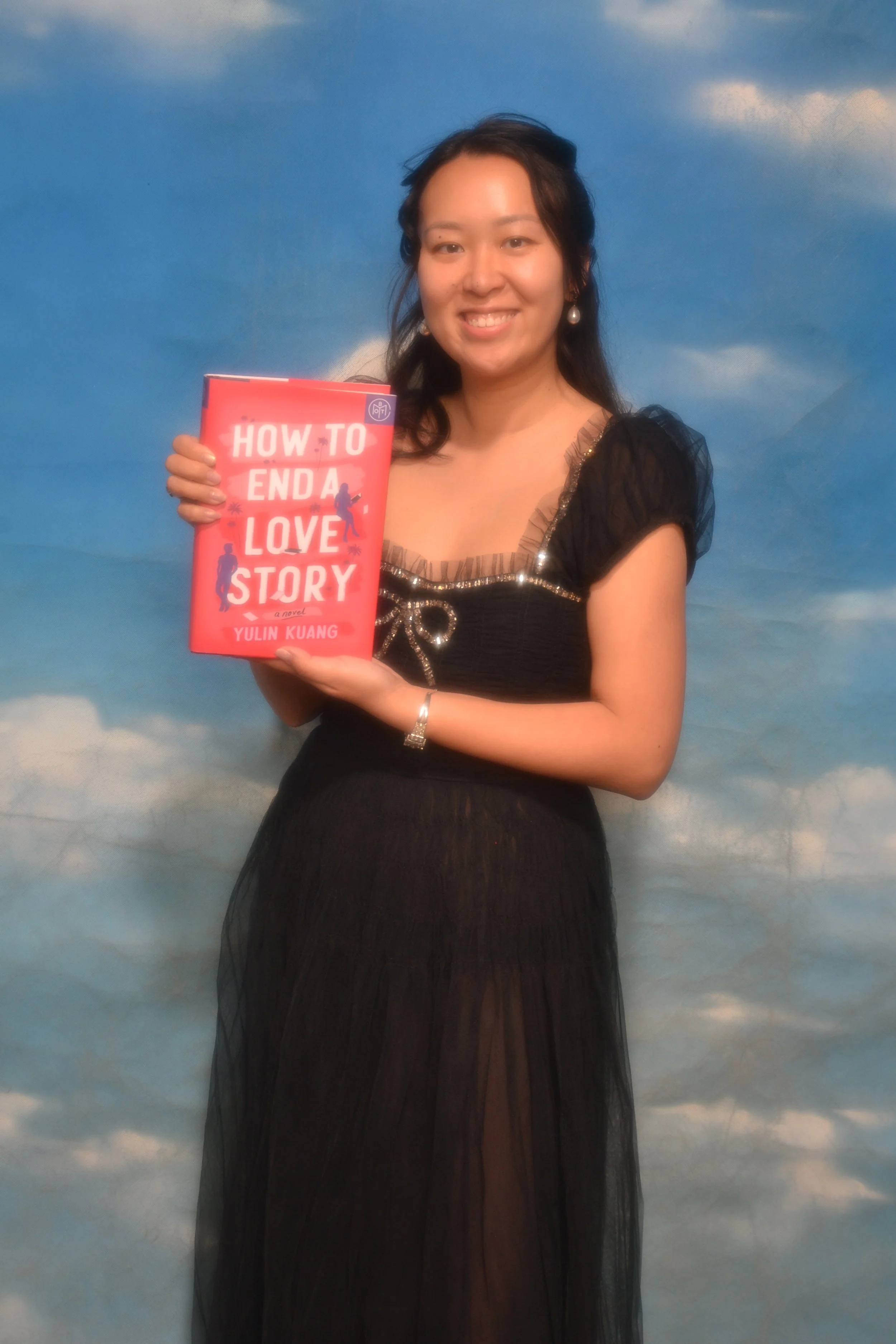 A woman in a black dress holding a book titled 'How to End a Love Story' by Yulin Kuang, standing in front of a sky backdrop.
