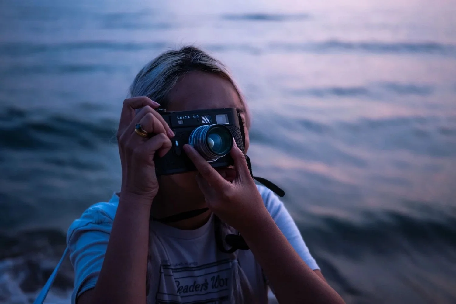 A woman with blonde hair taking a photograph with a vintage Leica camera near a body of water during dusk.