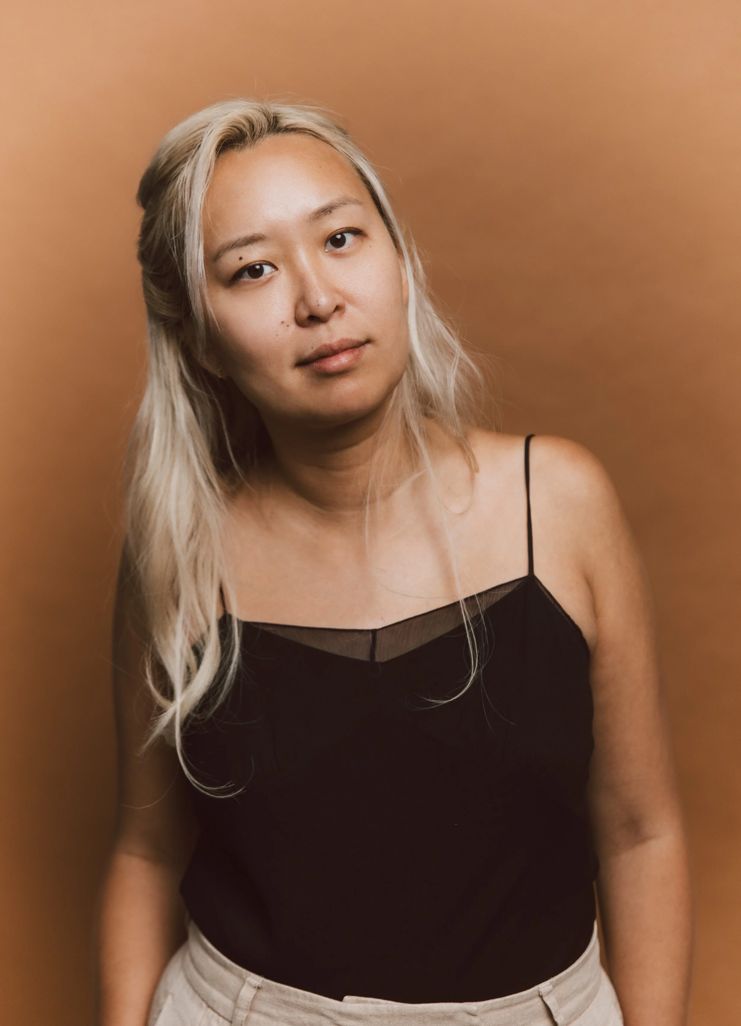 A young woman with long, wavy platinum blonde hair, wearing a black spaghetti strap top with a sheer neckline and beige pants, standing against a warm, tan-colored background.