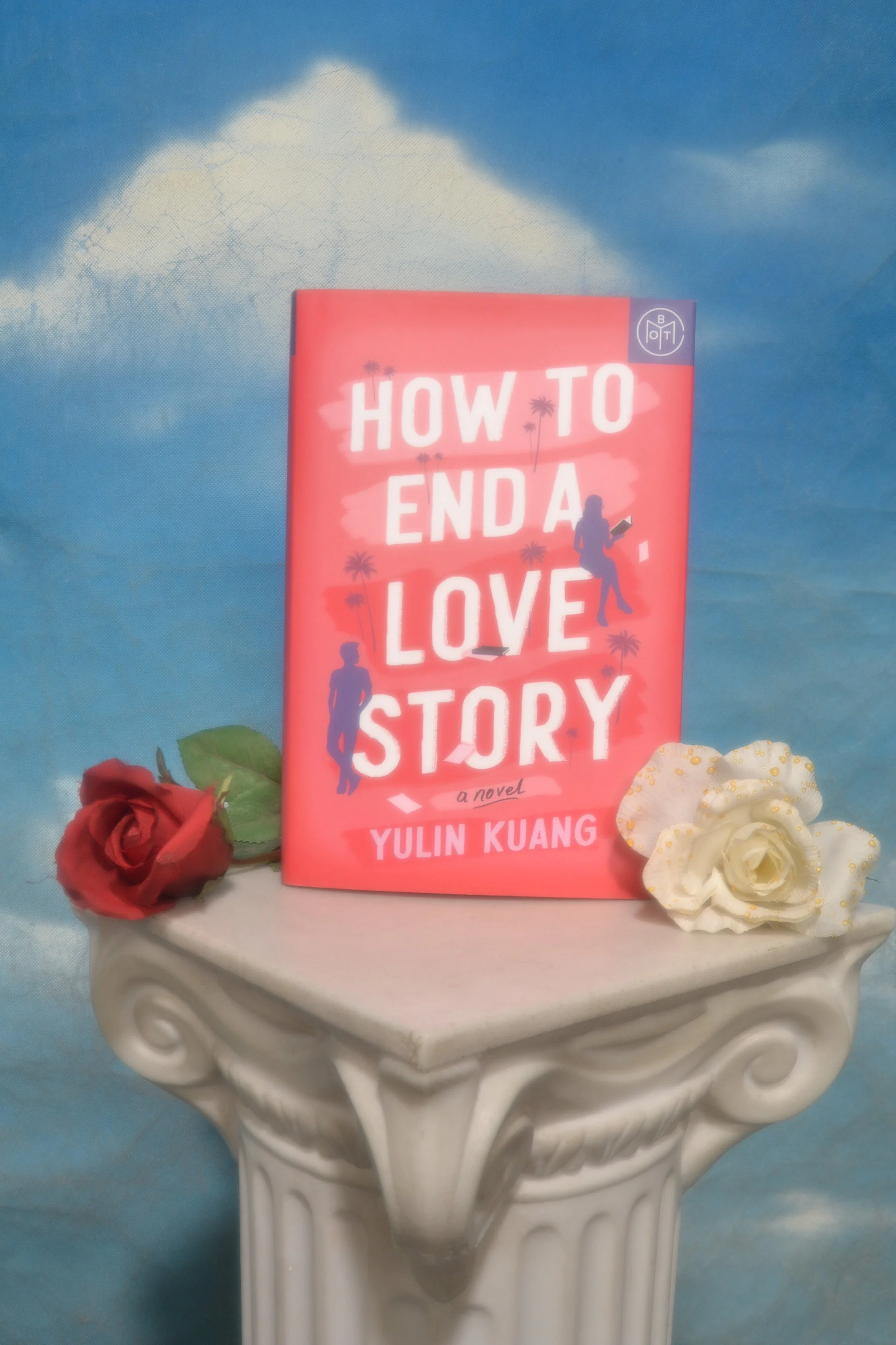 A pink book titled "How to End a Love Story" by Yulin Kuang is placed on a white decorative pedestal. There are two artificial roses, one red and one white, positioned on each side of the book. The background features a cloudy sky painted in blue and white.