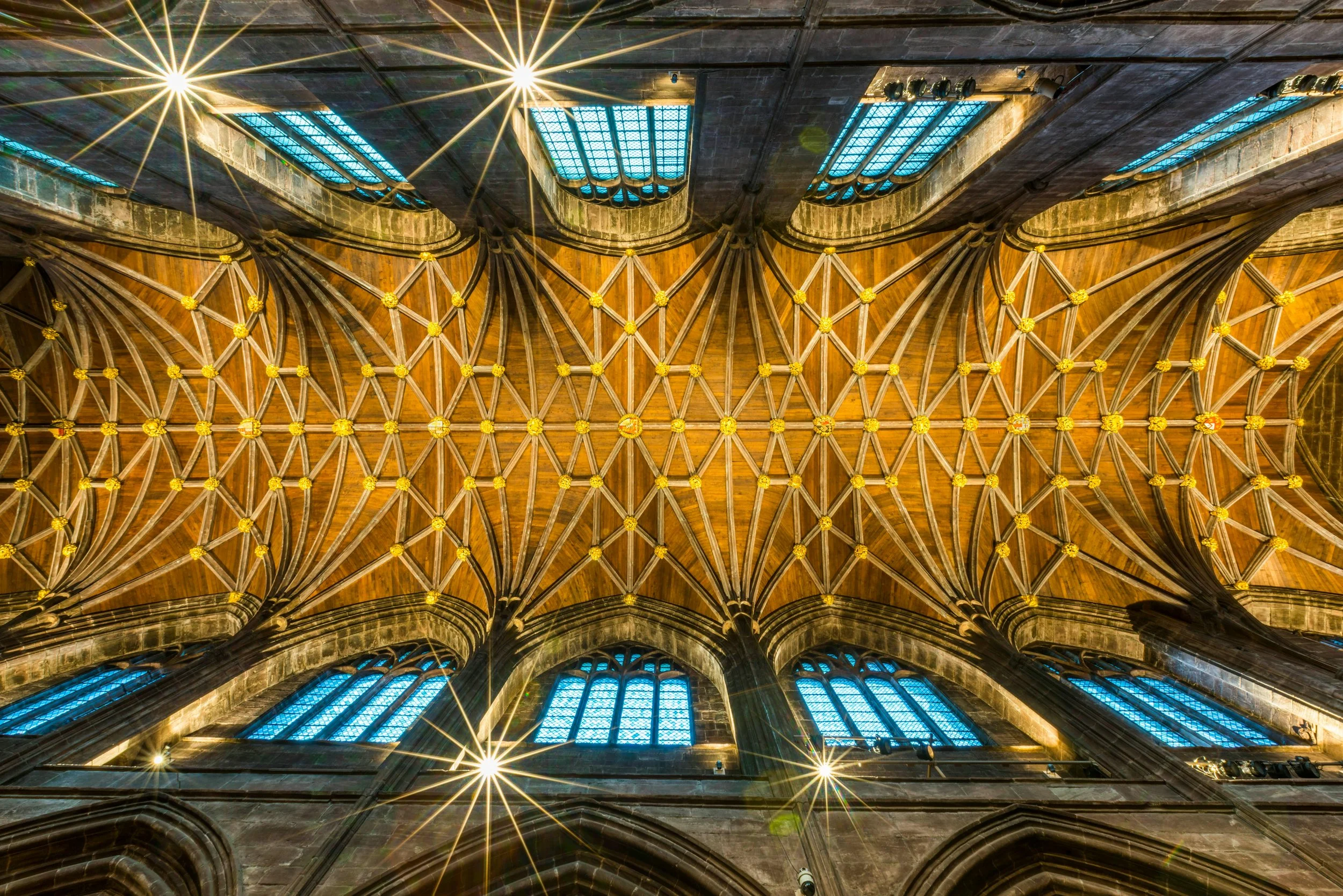 Looking up at the ceiling of a Gothic cathedral, showing intricate woodwork and stained glass windows shining sunlight.