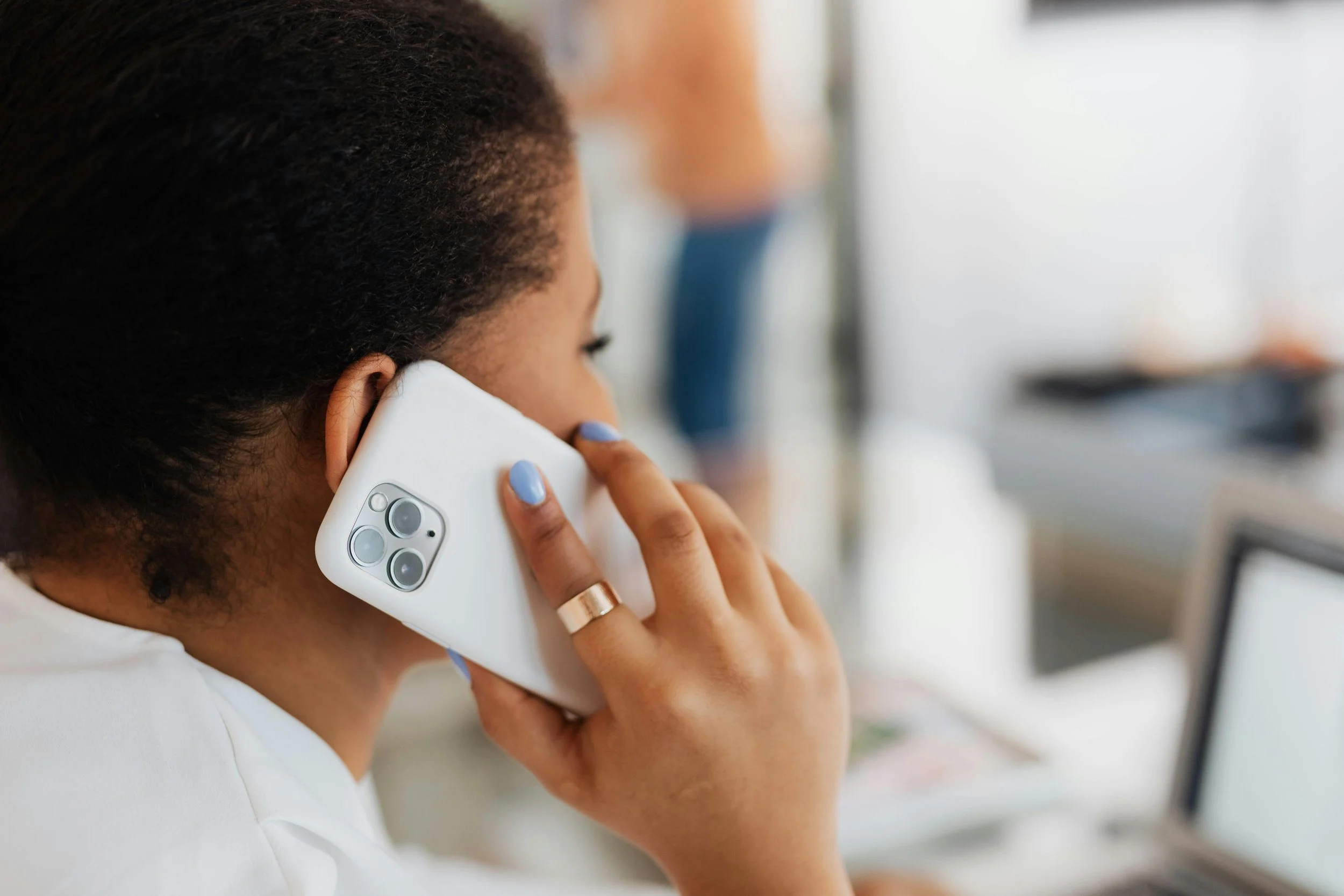 Woman with curly hair talking on a white smartphone, sitting at a desk with a laptop.