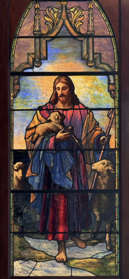 Stained glass window depicting Jesus Christ holding a lamb, with two sheep at his feet, set against a colorful sky background.