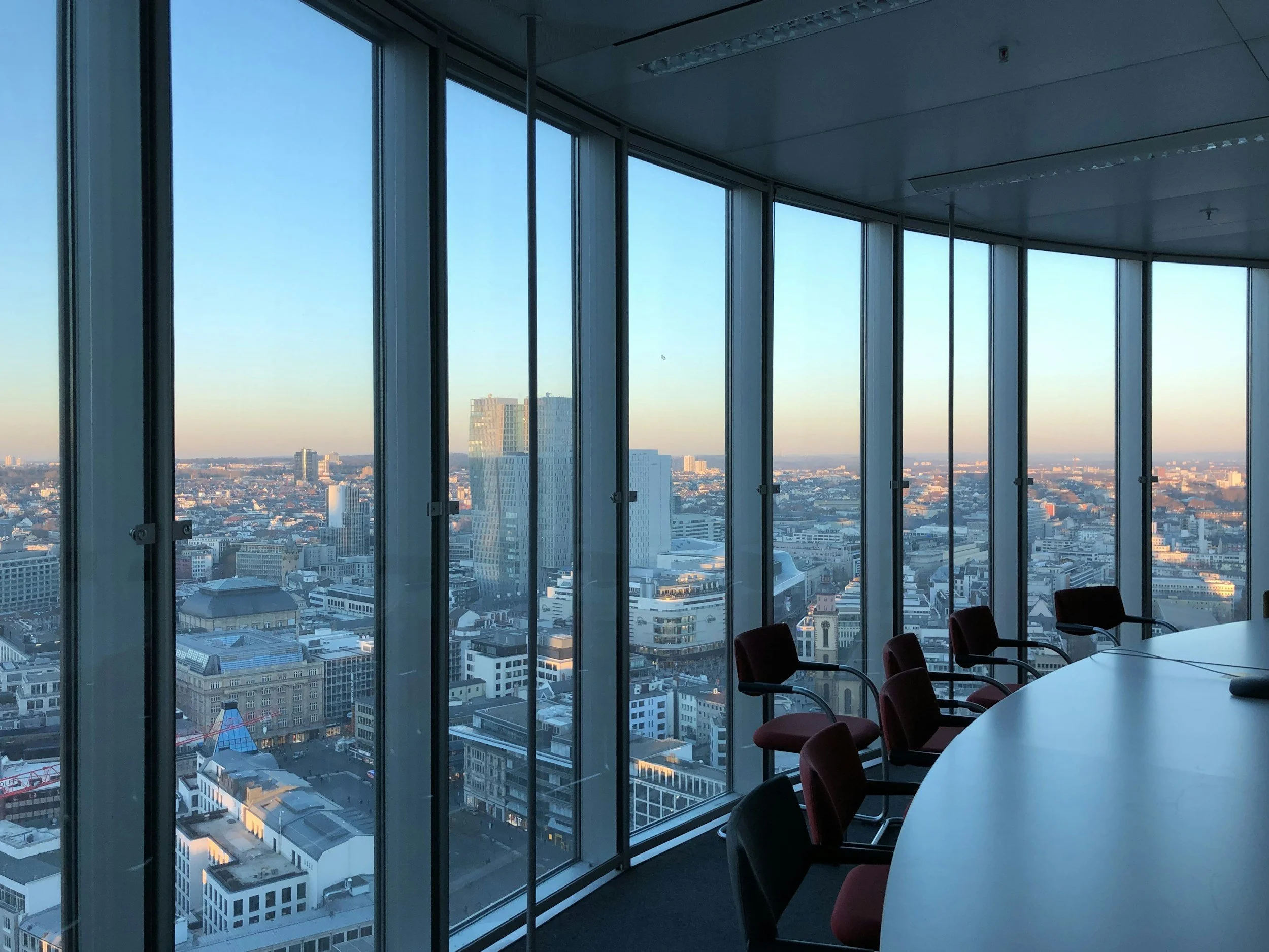 Interior of a high-rise conference room with large floor-to-ceiling windows showing a cityscape view during sunset, with a long rounded table and red chairs.