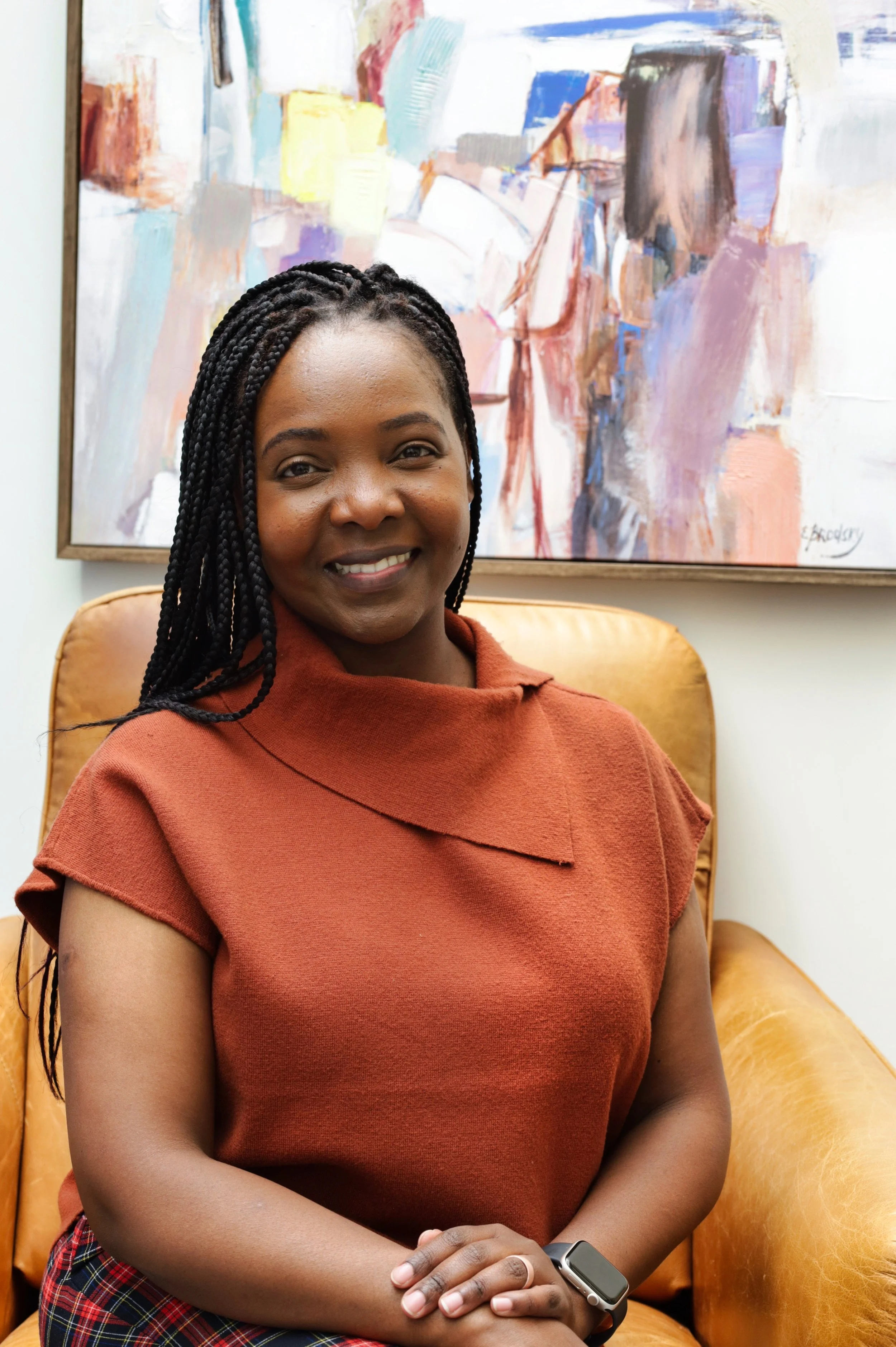 A woman with braided hair sitting on a tan leather chair in front of an abstract painting with colorful brushstrokes, smiling and wearing a burnt orange top and a smartwatch.