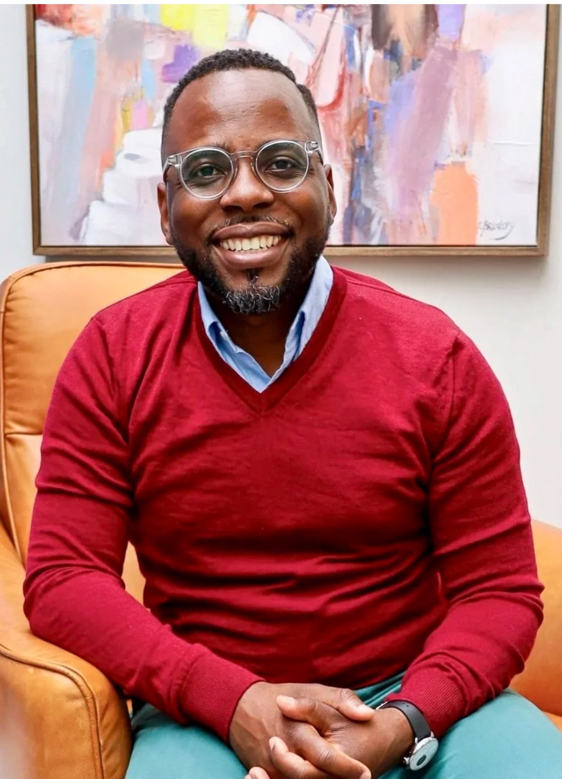 A smiling man wearing glasses, a red sweater over a collared blue shirt, and a black watch, sitting on an orange armchair in front of an abstract painting.