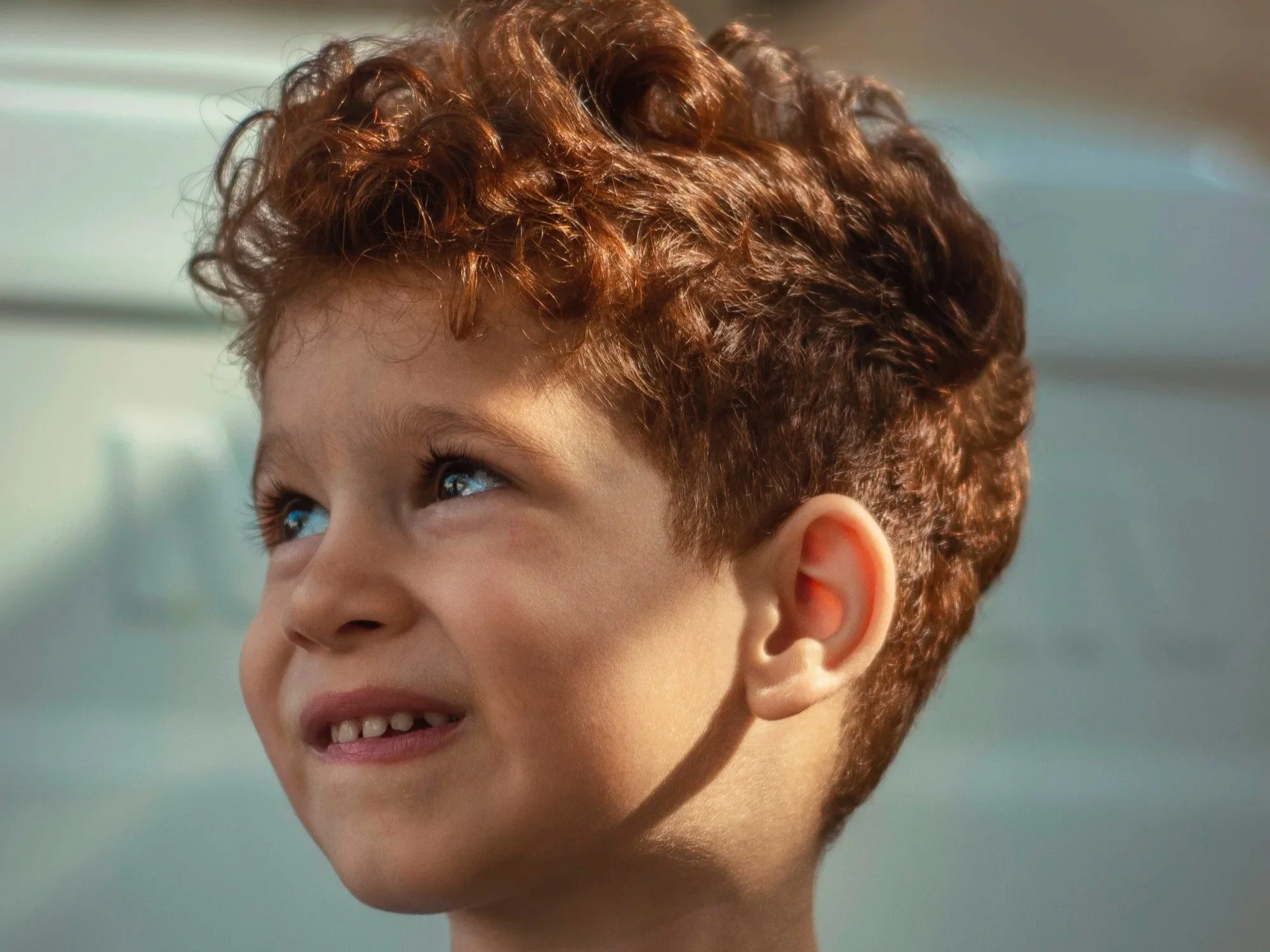 Close-up of a young boy with curly red hair looking upwards with a smile, indoors with natural light.
