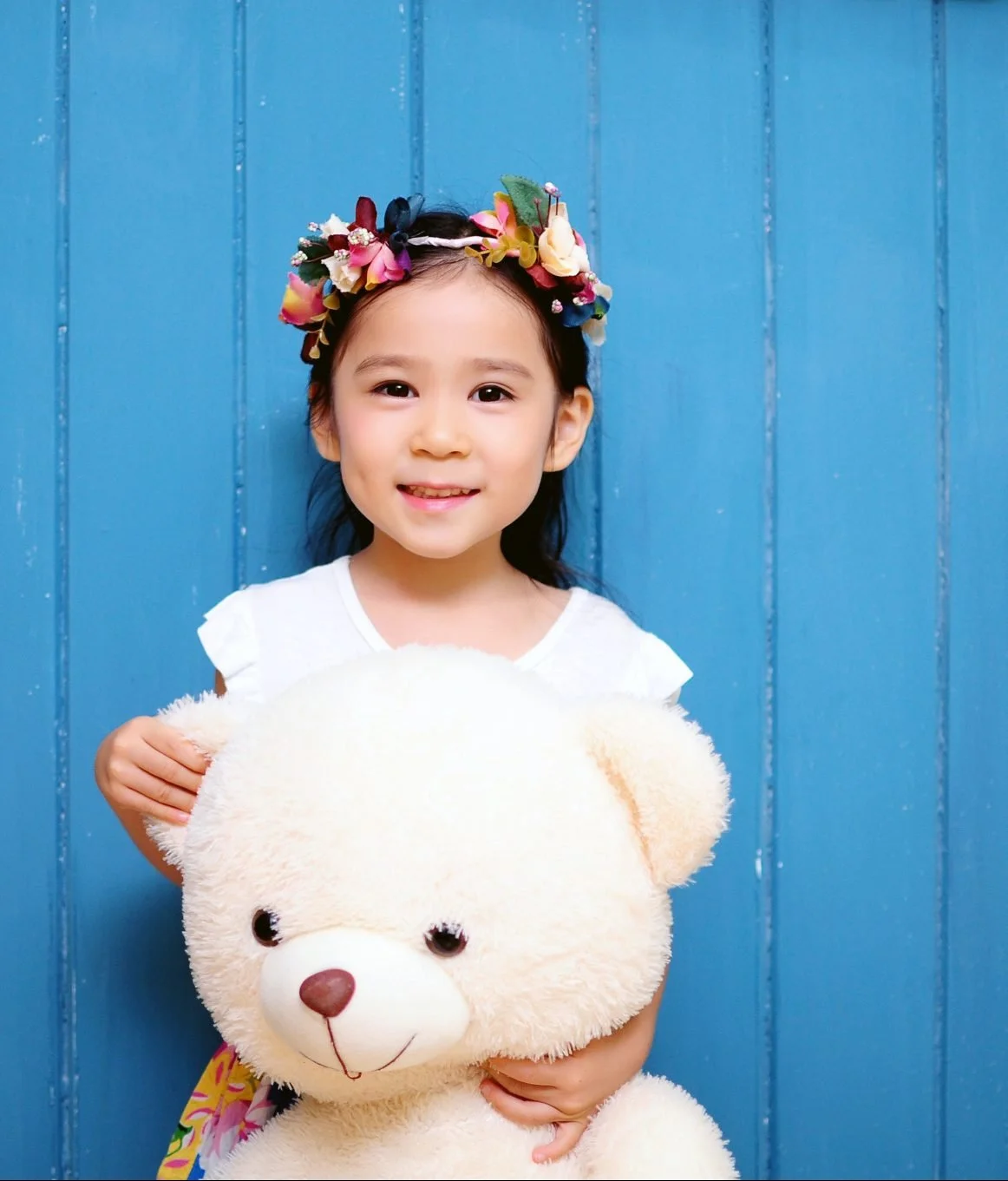 A young girl with a colorful flower crown smiling, holding a white teddy bear in front of a blue wooden background.