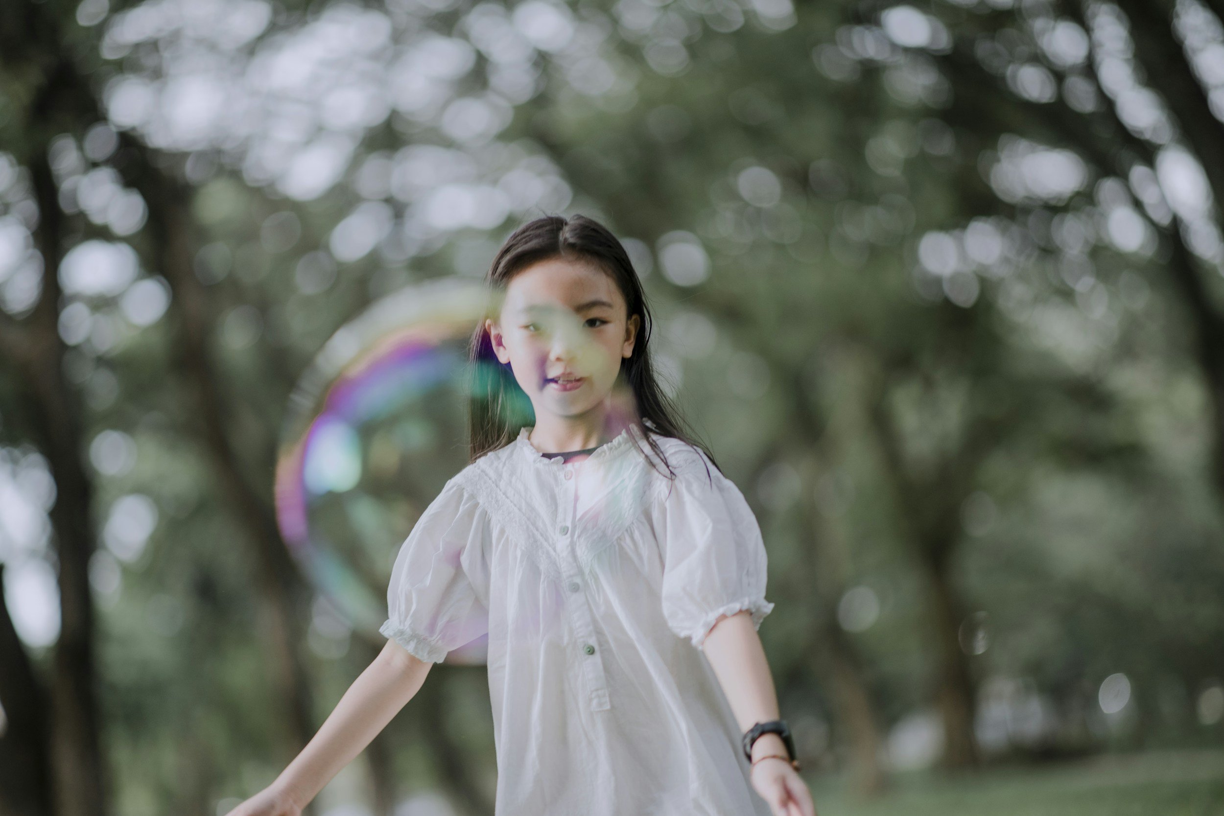 A young girl in a white dress is playing with a rainbow-colored soap bubble outdoors in a wooded area with green trees.