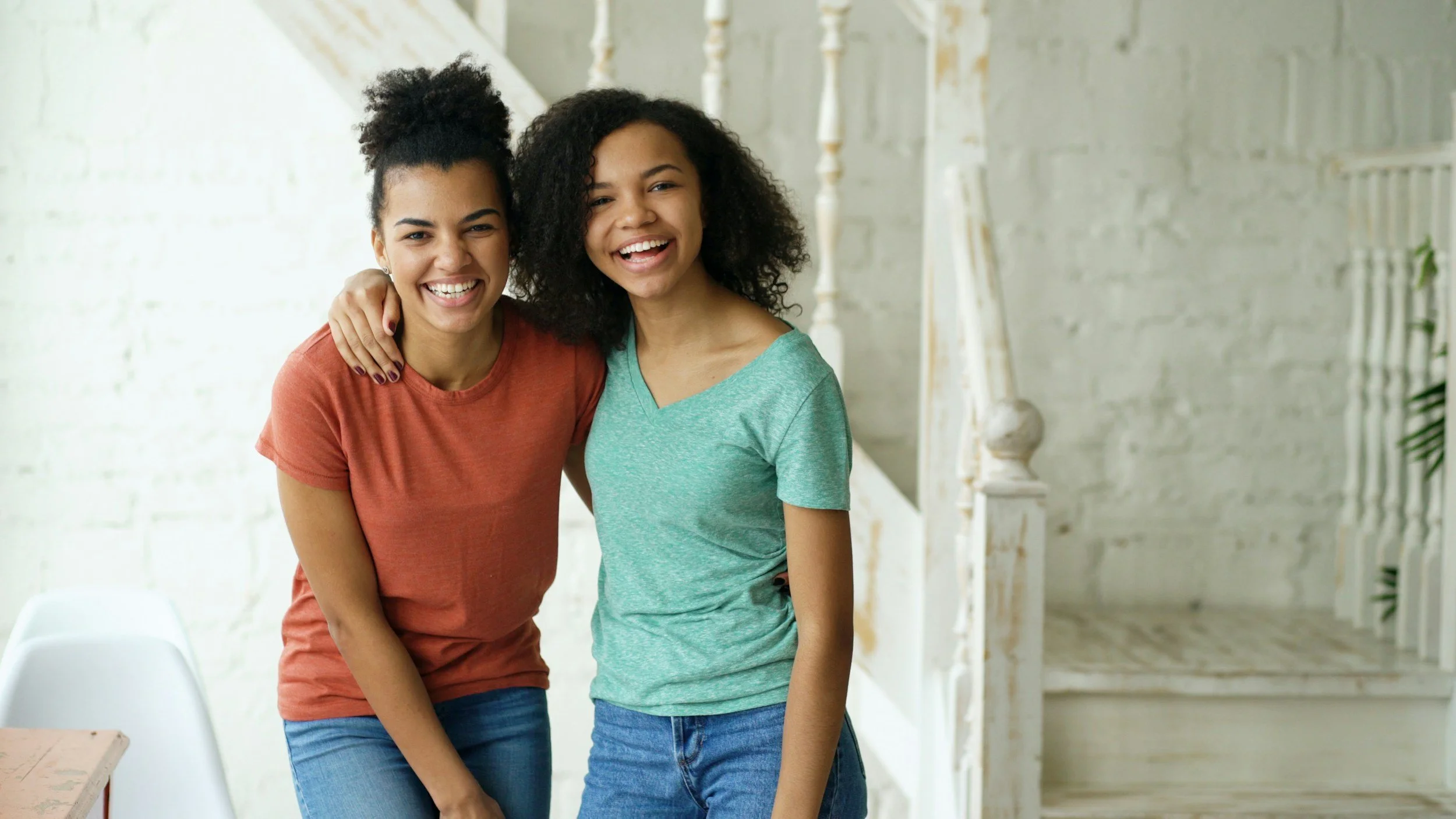 Two young women smiling and hugging each other indoors, standing near a staircase and a white wall.