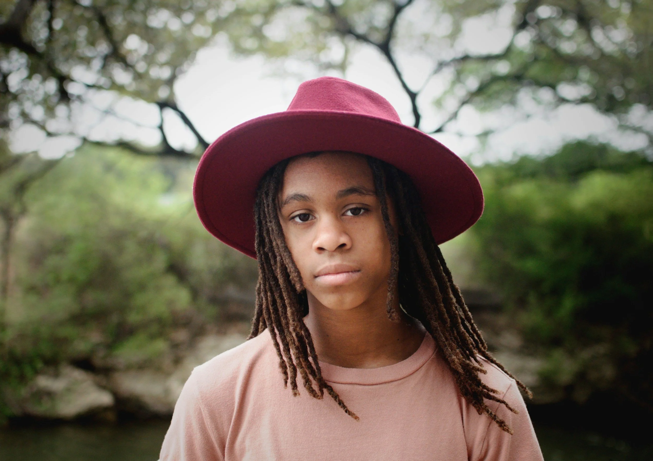 Young person with dreadlocks wearing a pink hat and a pink T-shirt outdoors in a green natural setting.