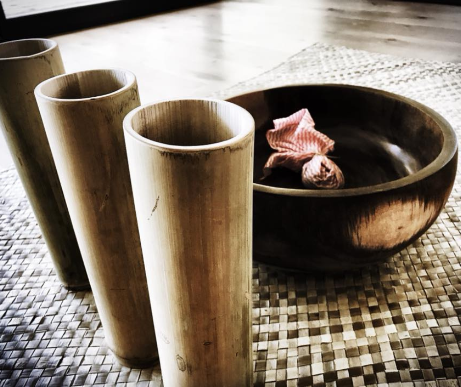 Three bamboo cups and a wooden bowl with a pink cloth inside, placed on a woven mat.