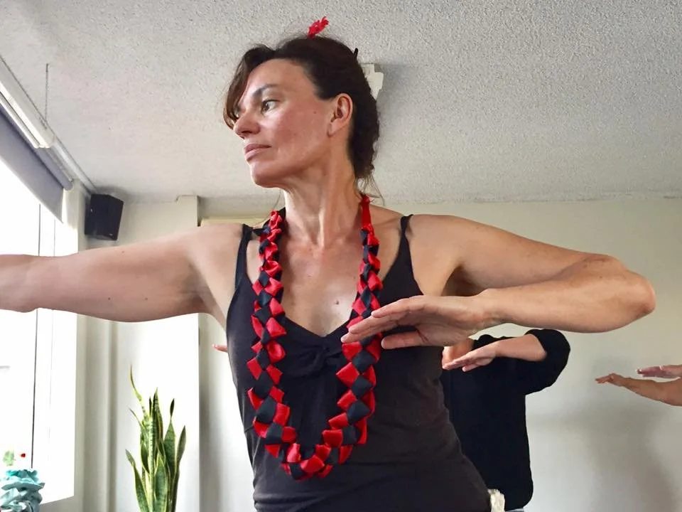 A woman in a black tank top participating in a fitness or dance class, with arms extended and wearing a red and black beaded necklace, in an indoor setting with a plant and other participants in the background.