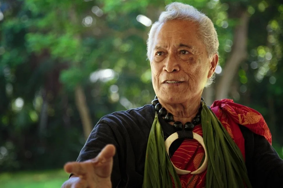 An elderly man with white hair, wearing traditional attire with green leaves, a beaded necklace, and a patterned red and black garment, standing outdoors with greenery in the background.