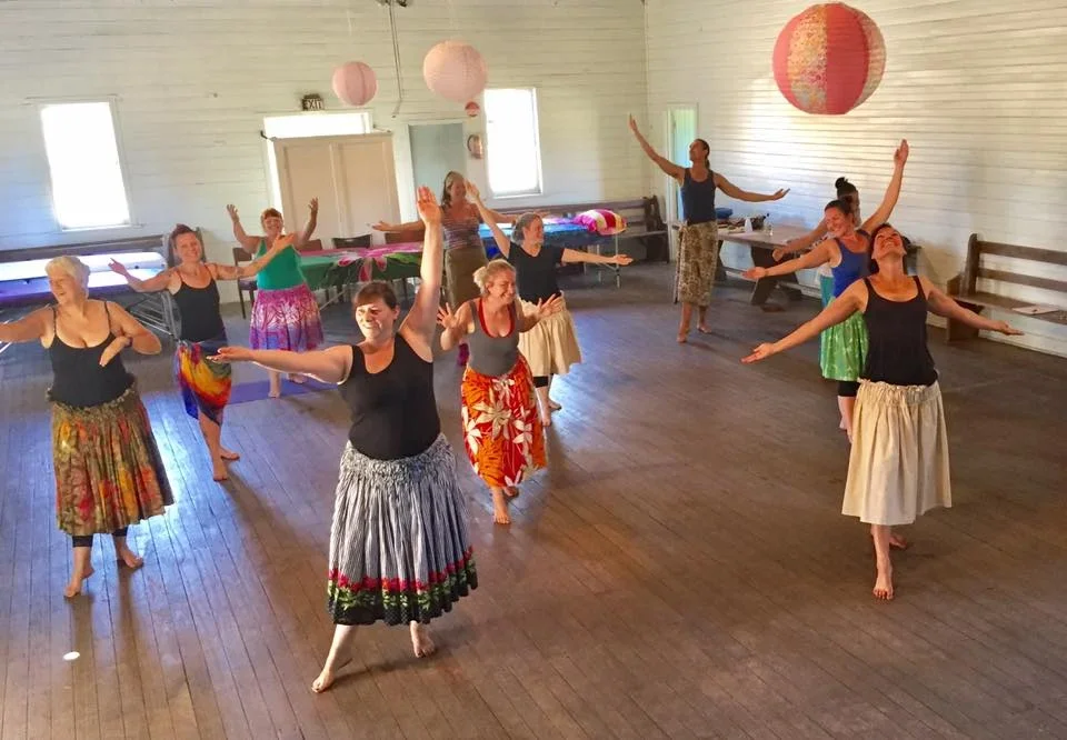 A group of women in colorful skirts practicing a dance in a spacious, well-lit room with wooden floors, paper lantern decorations, and tables along the walls.