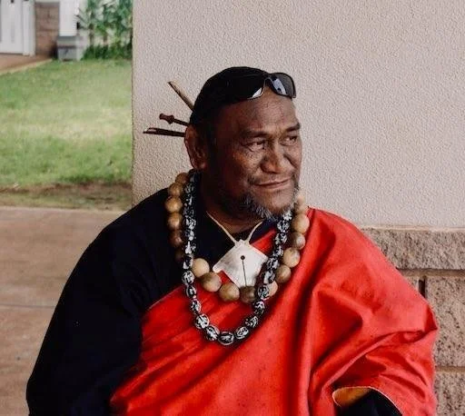 Elderly man wearing traditional Maasai attire, including a red shuka, multiple necklaces, and sunglasses on his head, sitting outdoors.