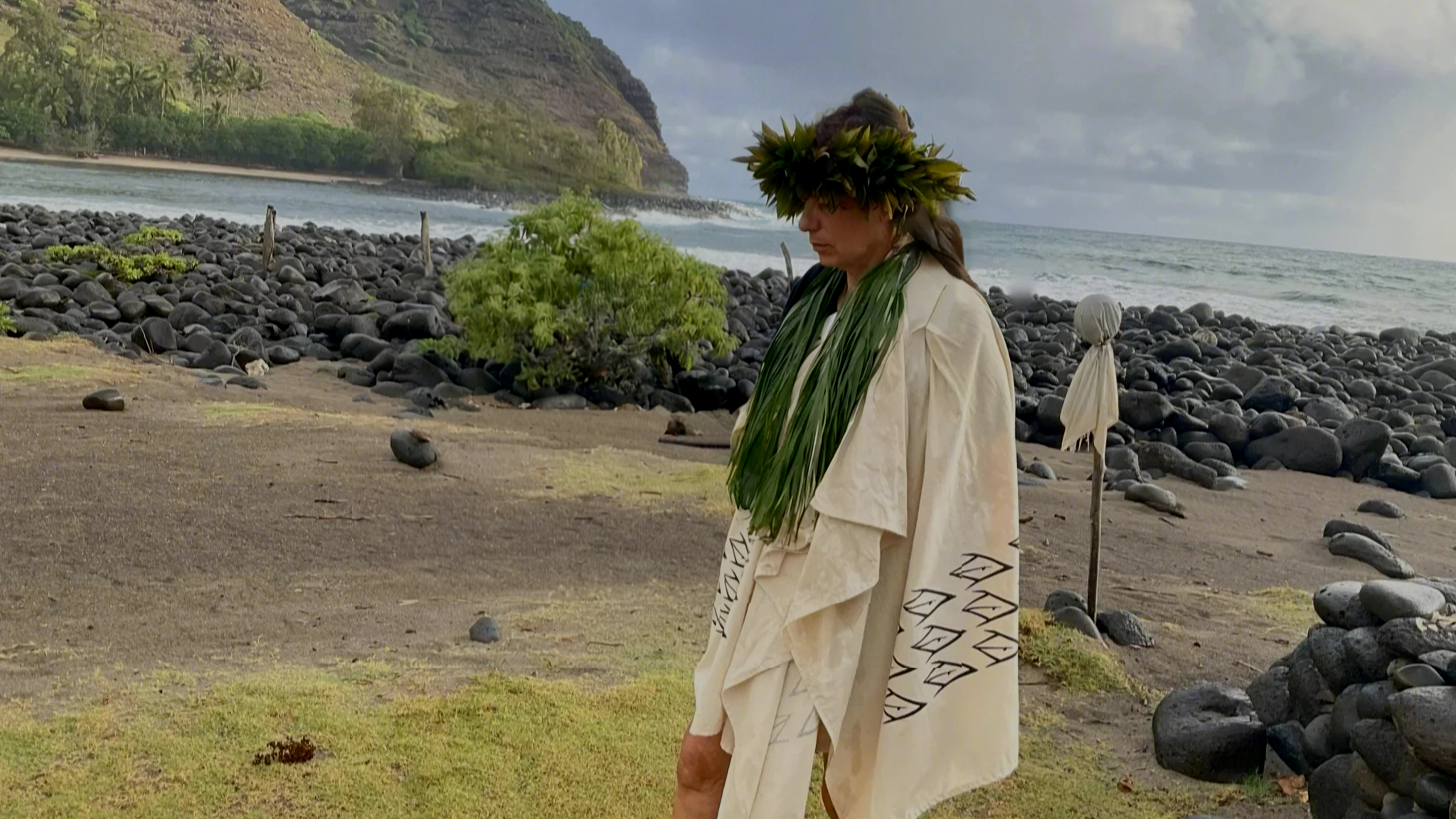 A woman dressed in traditional Polynesian attire, including a leafy crown and a white garment with black patterns, standing on a volcanic beach with black rocks, green vegetation, and a mountainous coastline in the background.